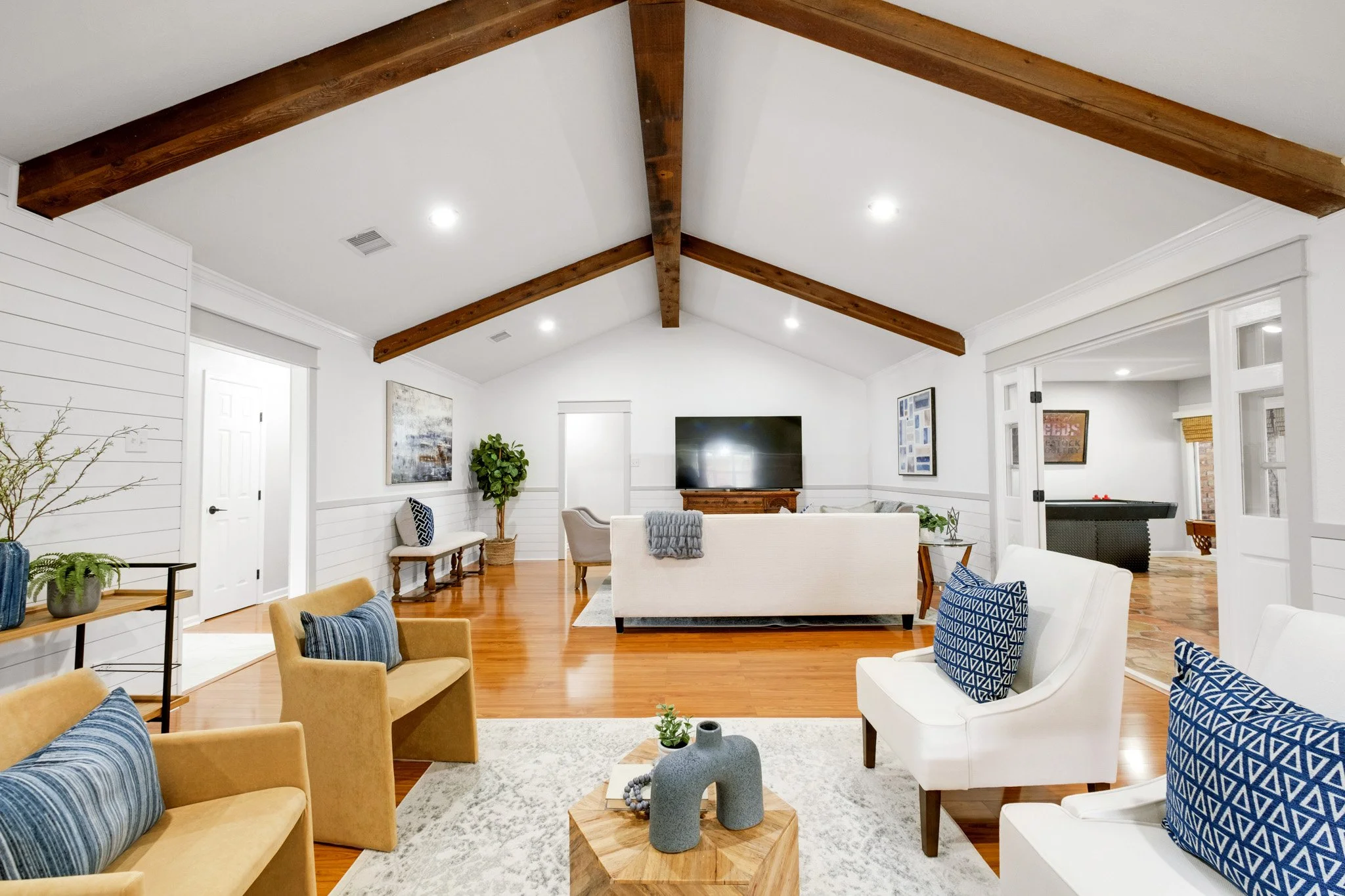 A spacious living room with white walls, wooden floor, and vaulted ceiling with exposed wooden beams. It is furnished with white and yellow armchairs, a white sofa with a gray throw, and a flat-screen TV on a wooden stand. Decor includes framed artwo