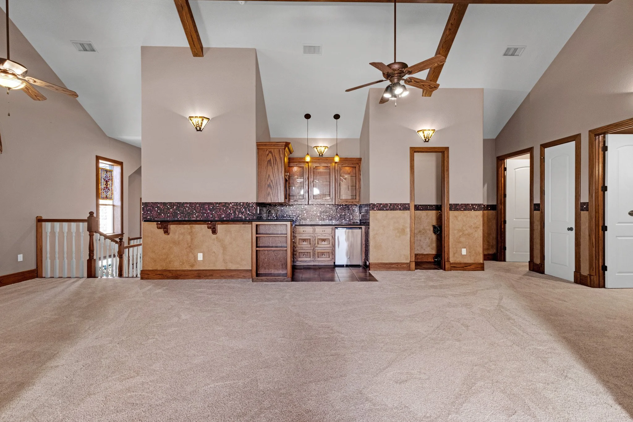 Open living space with beige carpet, wood accents, and a small kitchenette with upper and lower wooden cabinets, a small refrigerator, and a black granite countertop. Ceiling fans and wall-mounted lights are visible.