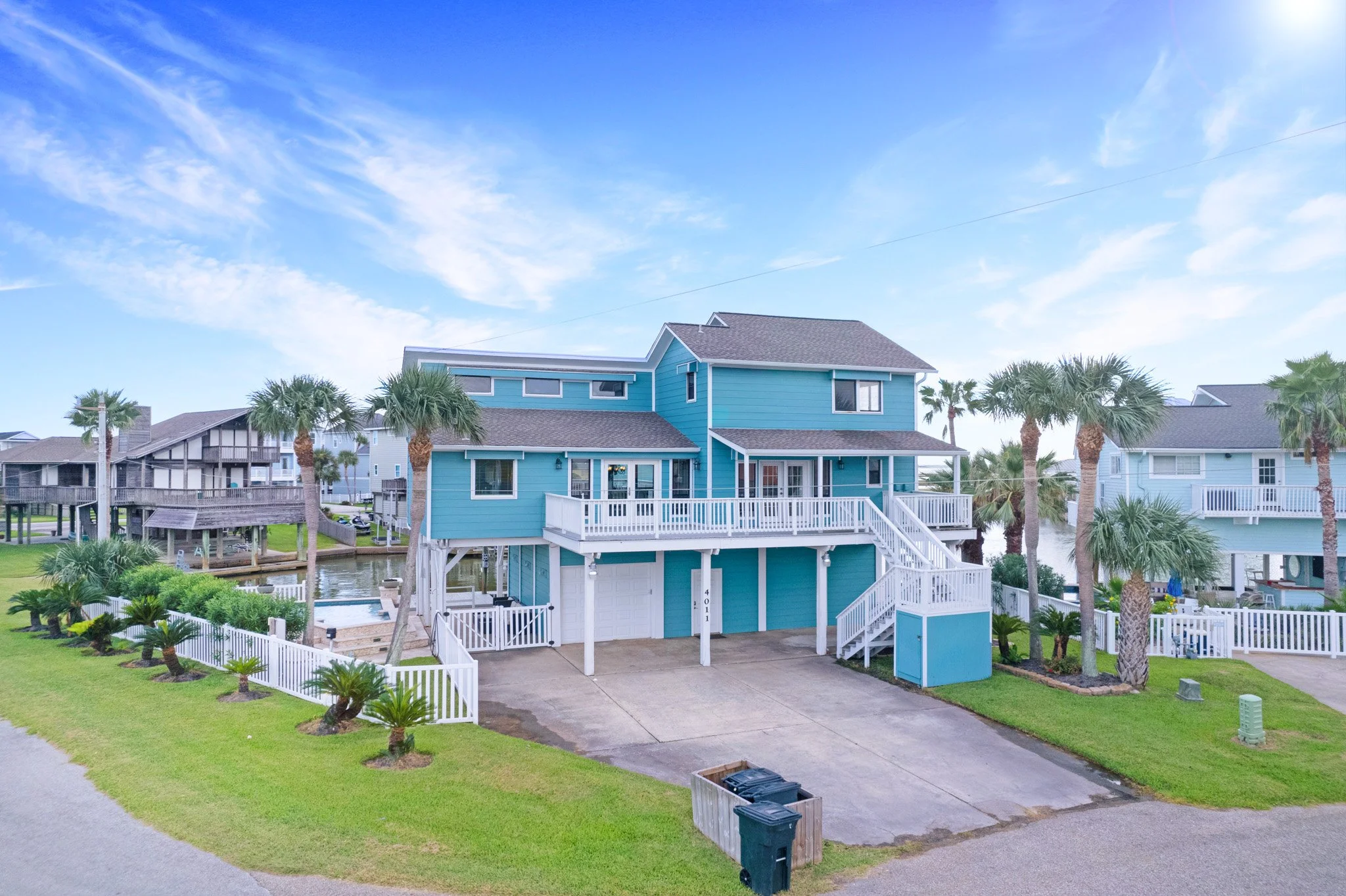 A two-story beach house painted in blue with balconies, surrounded by palm trees, located near a canal with neighboring homes in the background.