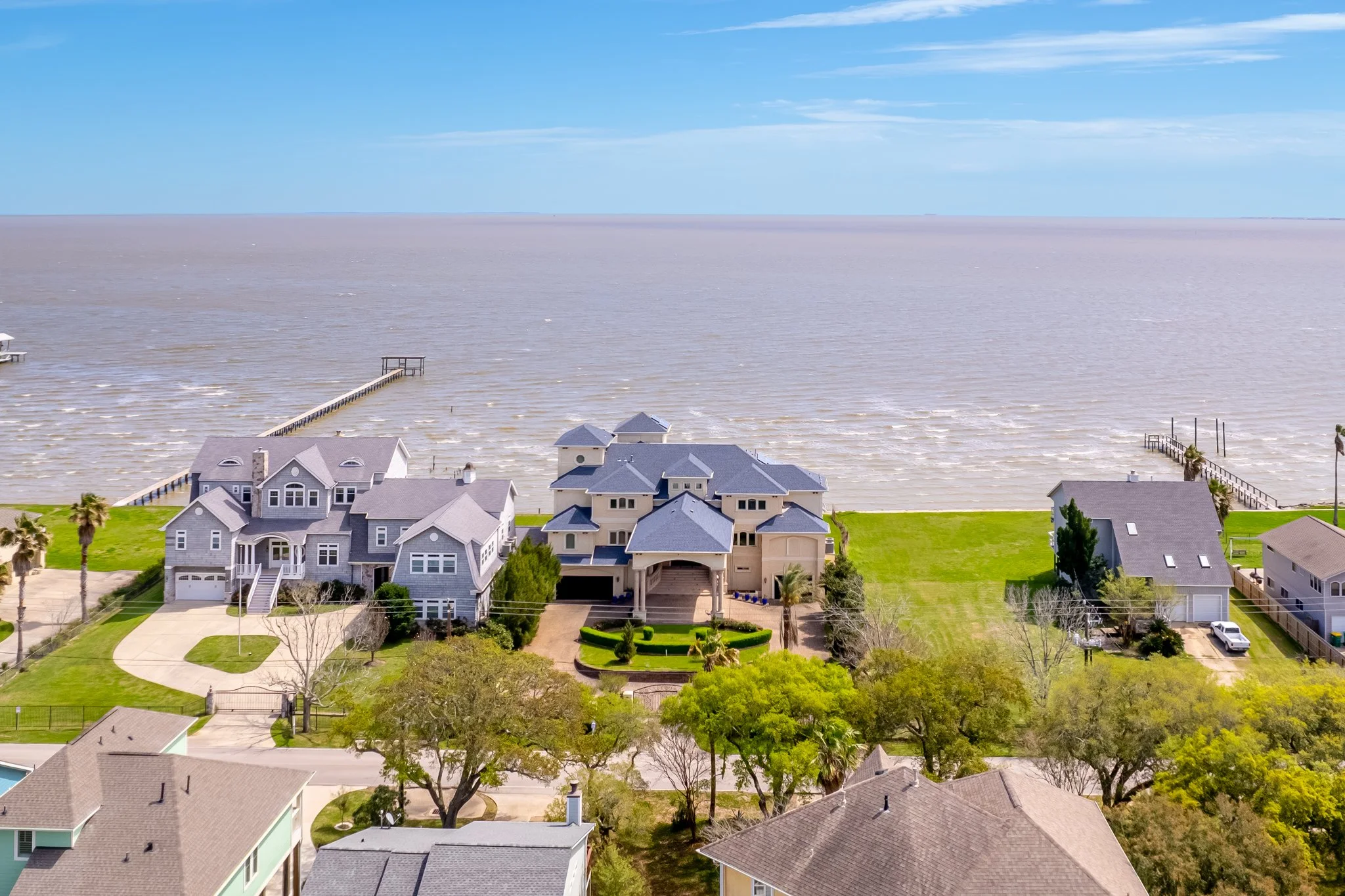 Aerial view of a beachfront neighborhood with large houses, including a prominent multi-story home with a blue roof, green lawns, trees, and a view of the ocean with piers extending into the water.