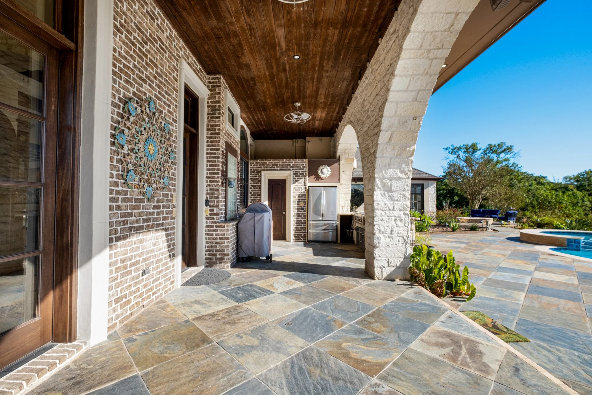 Backyard patio with brick and stone walls, tiled floor, outdoor kitchen, and pool area in the background under a clear blue sky.
