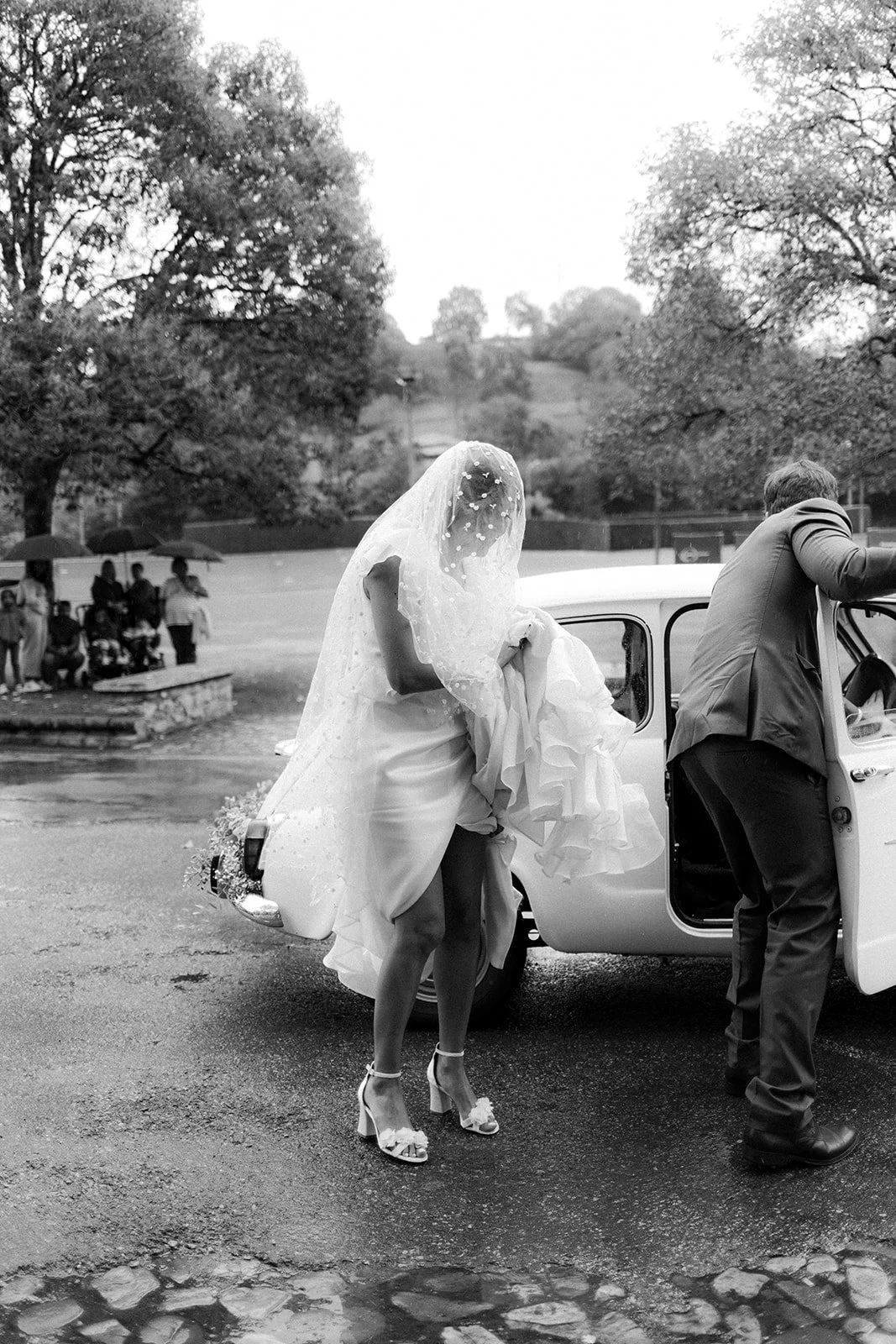 Novia subiendo a un coche clásico bajo la lluvia en una boda en el norte de España. Fotografía artística en blanco y negro que aprovecha el clima para crear una estética cinematográfica.