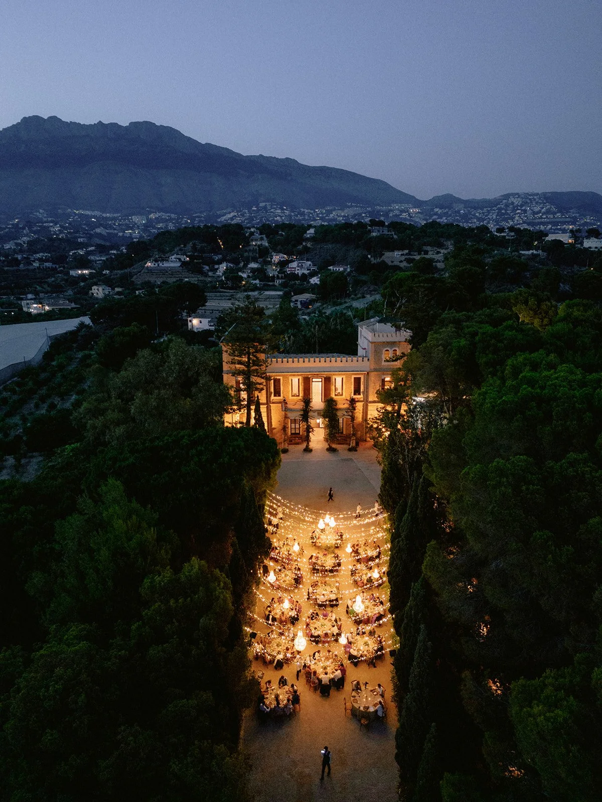 Vista aérea al anochecer de una cena de boda al aire libre iluminada con guirnaldas de luces en la finca Marqués de Montemolar, Altea. Ambiente sofisticado y romántico.