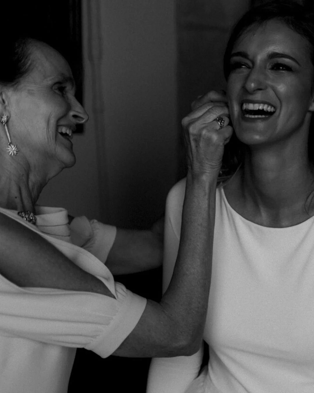 Madre ayudando a la novia a ponerse los pendientes durante los preparativos, riendo y disfrutando el momento. Fotografía documental que captura la conexión madre e hija.