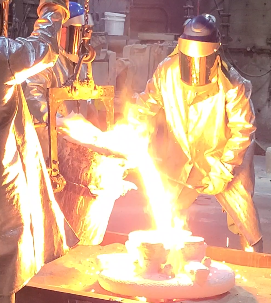 Molten metal being poured into a cast by two people in protective gear.