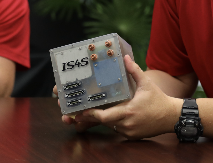 Over-the-should shot of a man holding a clear plastic cube with the IS4S logo on the side and that is apparently full of various hardware and wires.