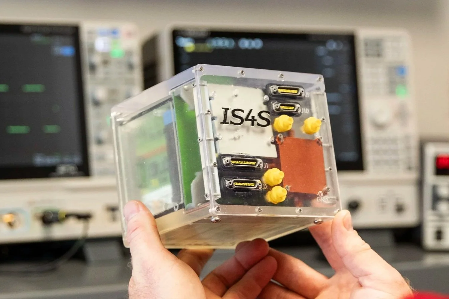 Over-the-should shot of a man holding a clear plastic cube with the IS4S logo on the side and that is apparently full of various hardware and wires.