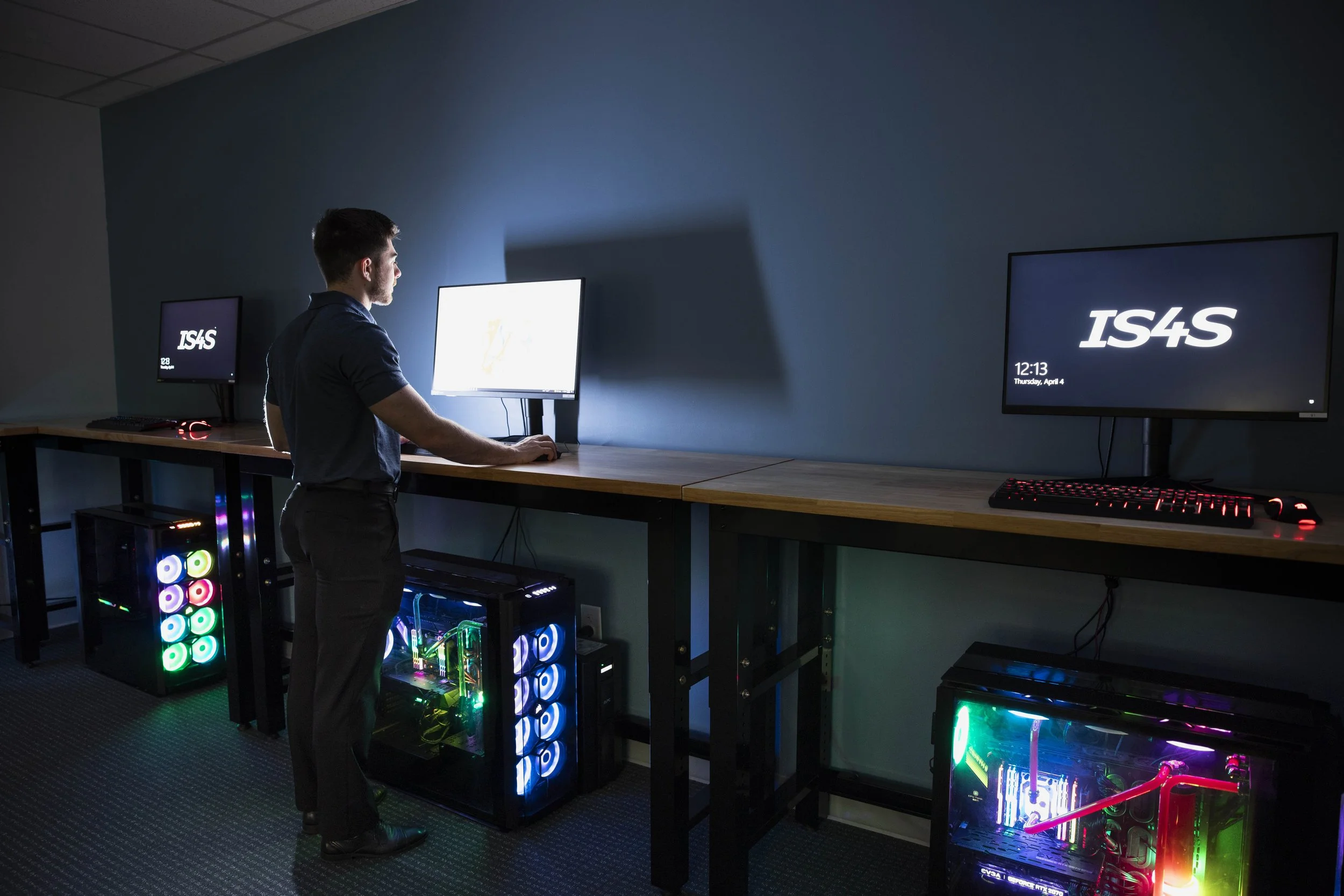 A man standing at a tall desk working on a computer monitor, under the desk are computer towers with multi-colored display lights.