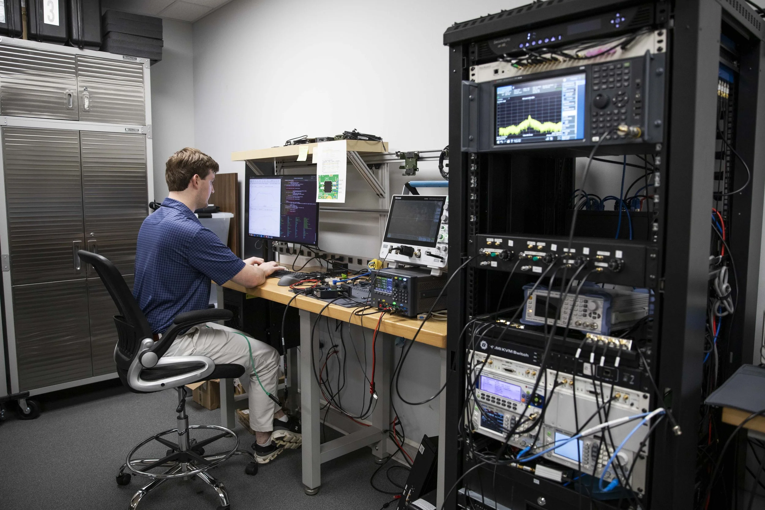 A man sitting at a desk next to a rack with a lot of technical equipment on it including a screen with a non-specific chart on it.
