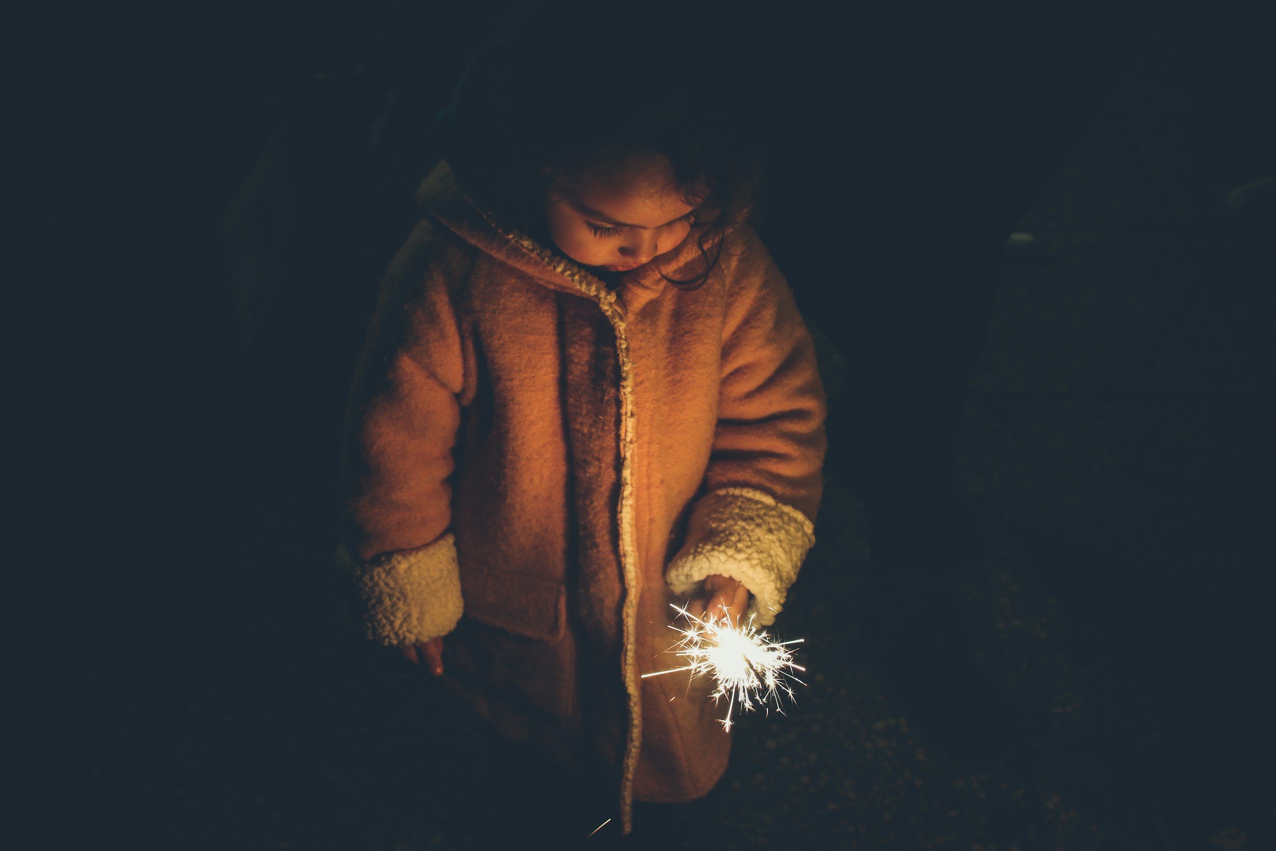 A young girl in a warm coat holding a lit sparkler during the night.