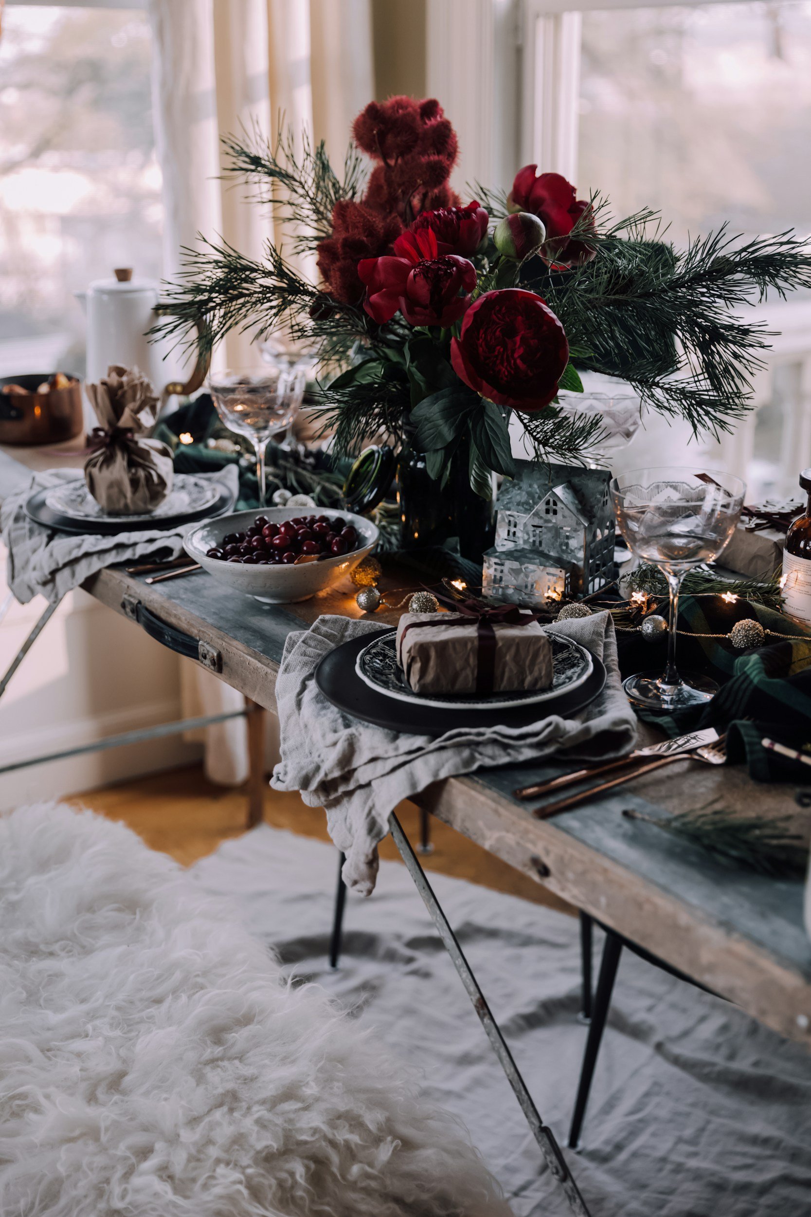 A rustic wooden dining table decorated with a large floral centerpiece featuring red flowers and greenery, set with black plates, wine glasses, wrapped gifts, candle holders, and string lights, in a cozy room with natural light.