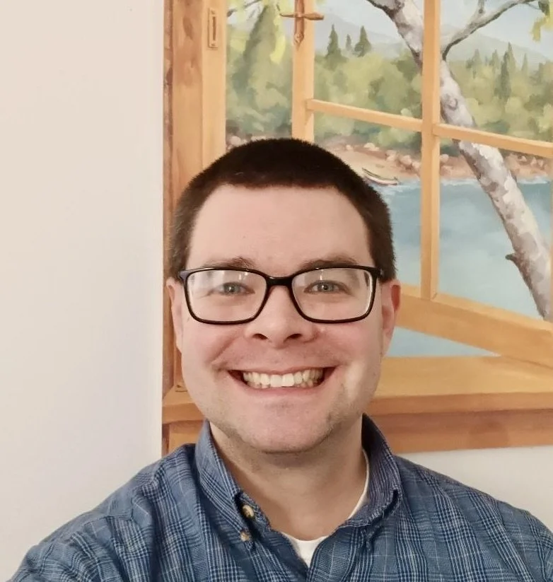 A man smiling and wearing glasses, in front of a window with a painted landscape of trees, water, and a shoreline.