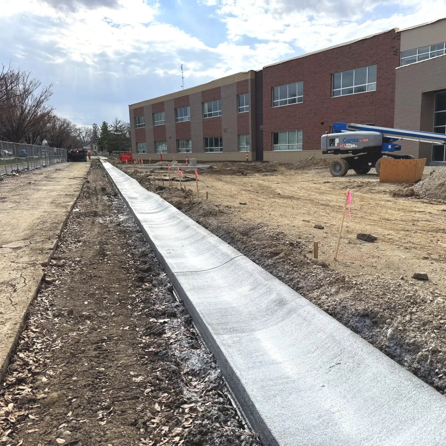 Construction site with sidewalk being paved, construction equipment, dirt piles, and a building in the background.