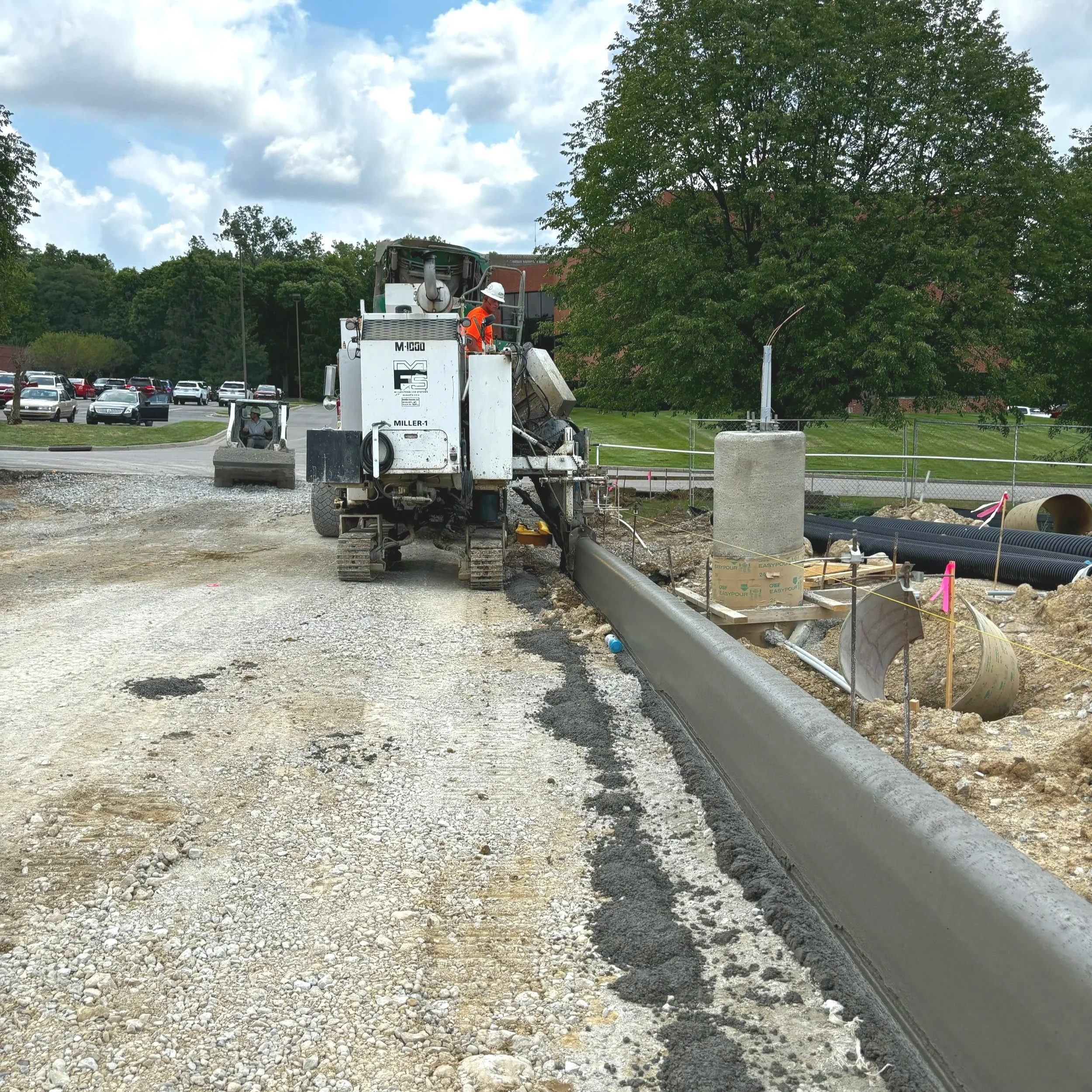 Construction worker operating heavy machinery on a road construction site with a large tree and parked cars in the background.