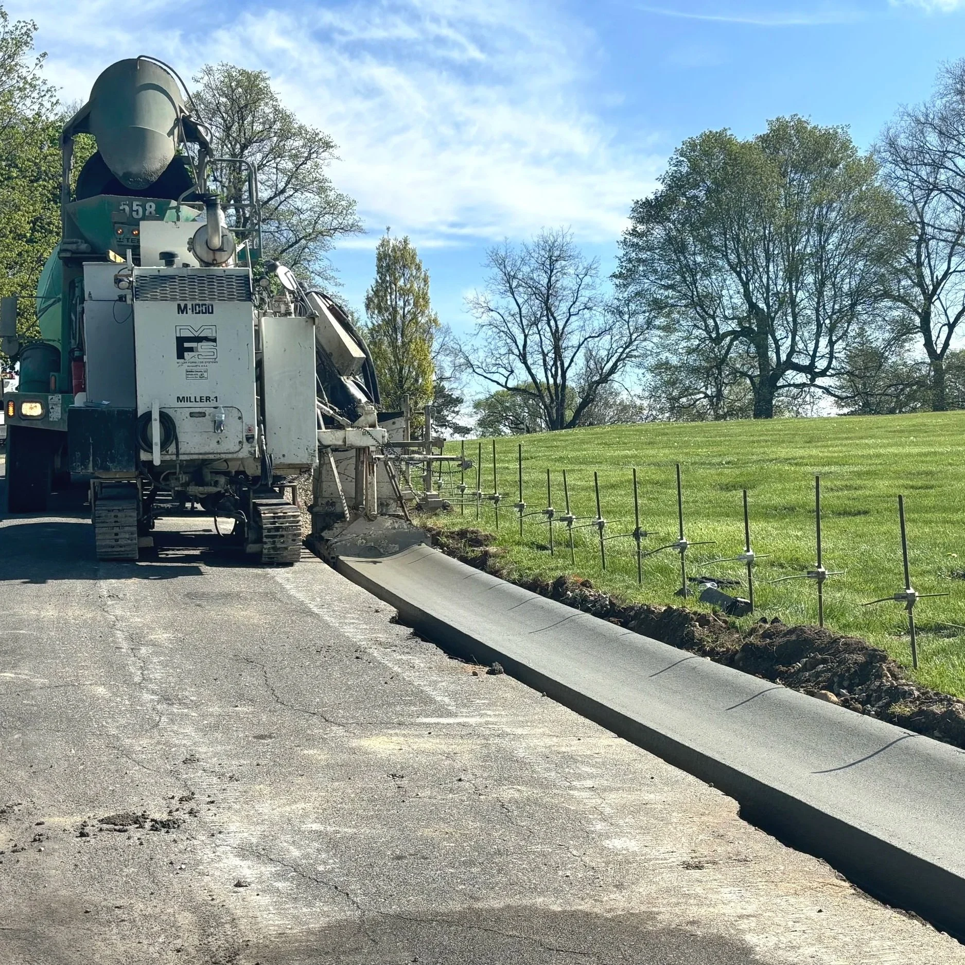 A road construction worker machine pouring fresh concrete along the edge of a new sidewalk or curb, with green grass and trees in the background.