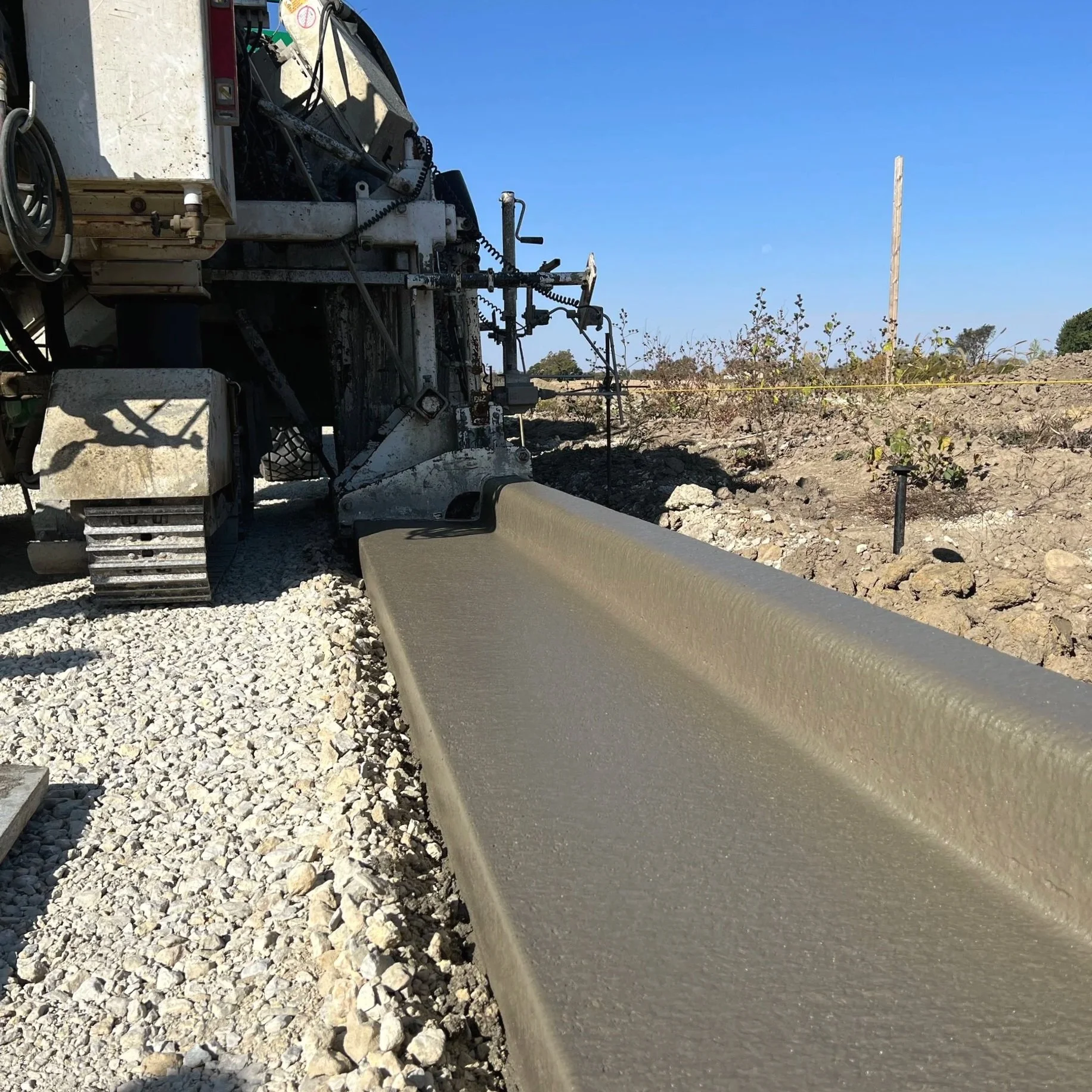 A construction site with freshly poured concrete on a long, narrow strip of land. Construction machinery is visible on the left side, and the background shows a clear blue sky with some trees and a utility pole.