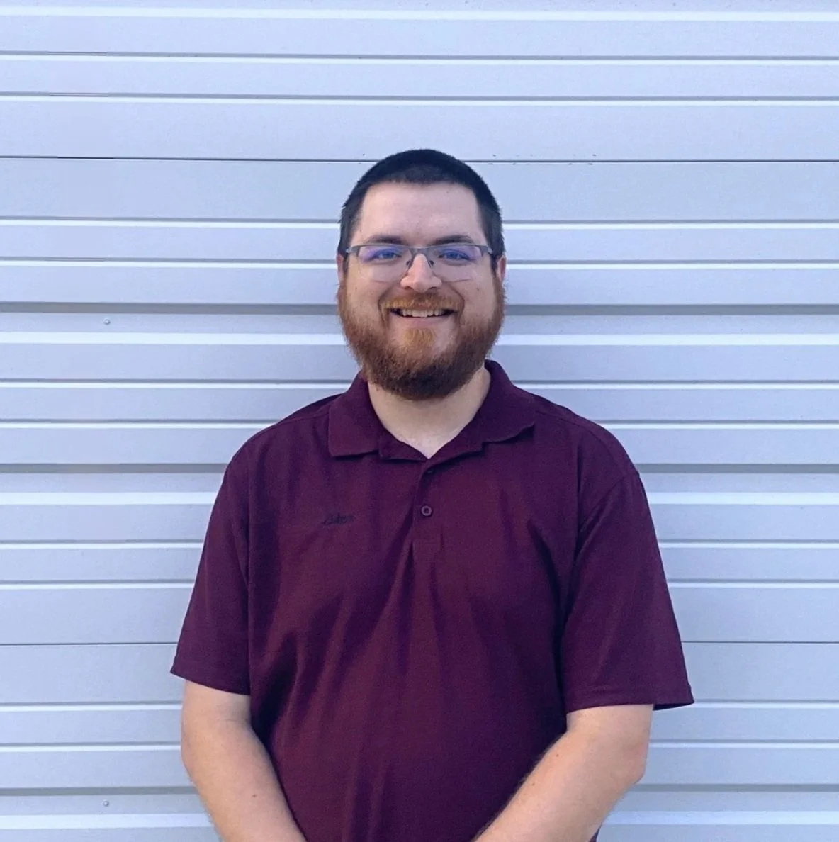 A man with a red beard and short brown hair smiling, wearing a brown and navy plaid shirt, standing against a plain white wall.