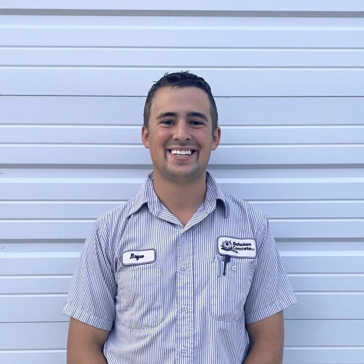 A young man with short brown hair, wearing a plaid button-up shirt and an orange T-shirt, smiling against a plain off-white wall.