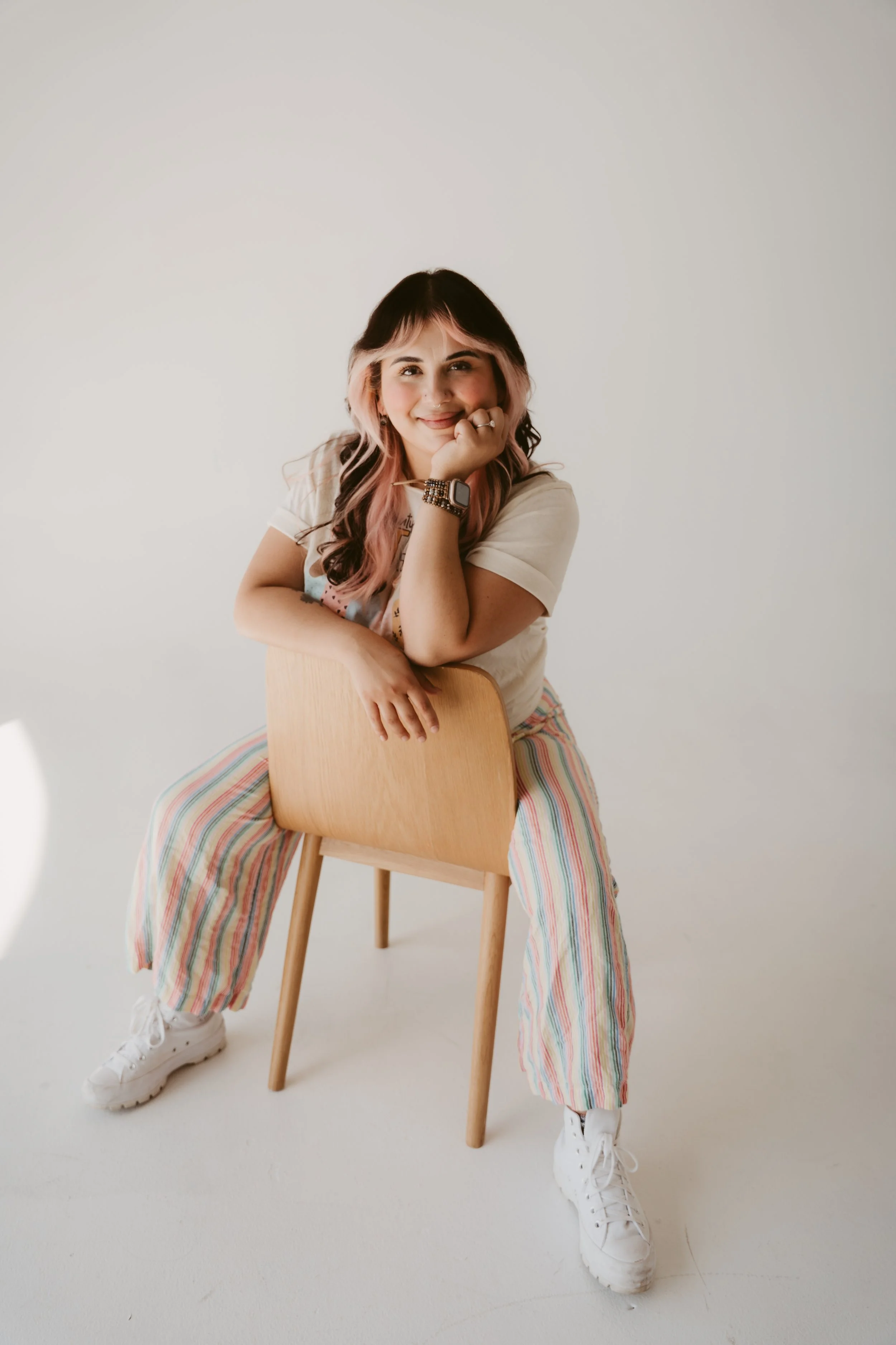 A young woman with pink and black hair sitting on a wooden chair against a plain white background, wearing a light-colored t-shirt, colorful striped wide-leg pants, white sneakers, and a watch, smiling and resting her chin on her hand.