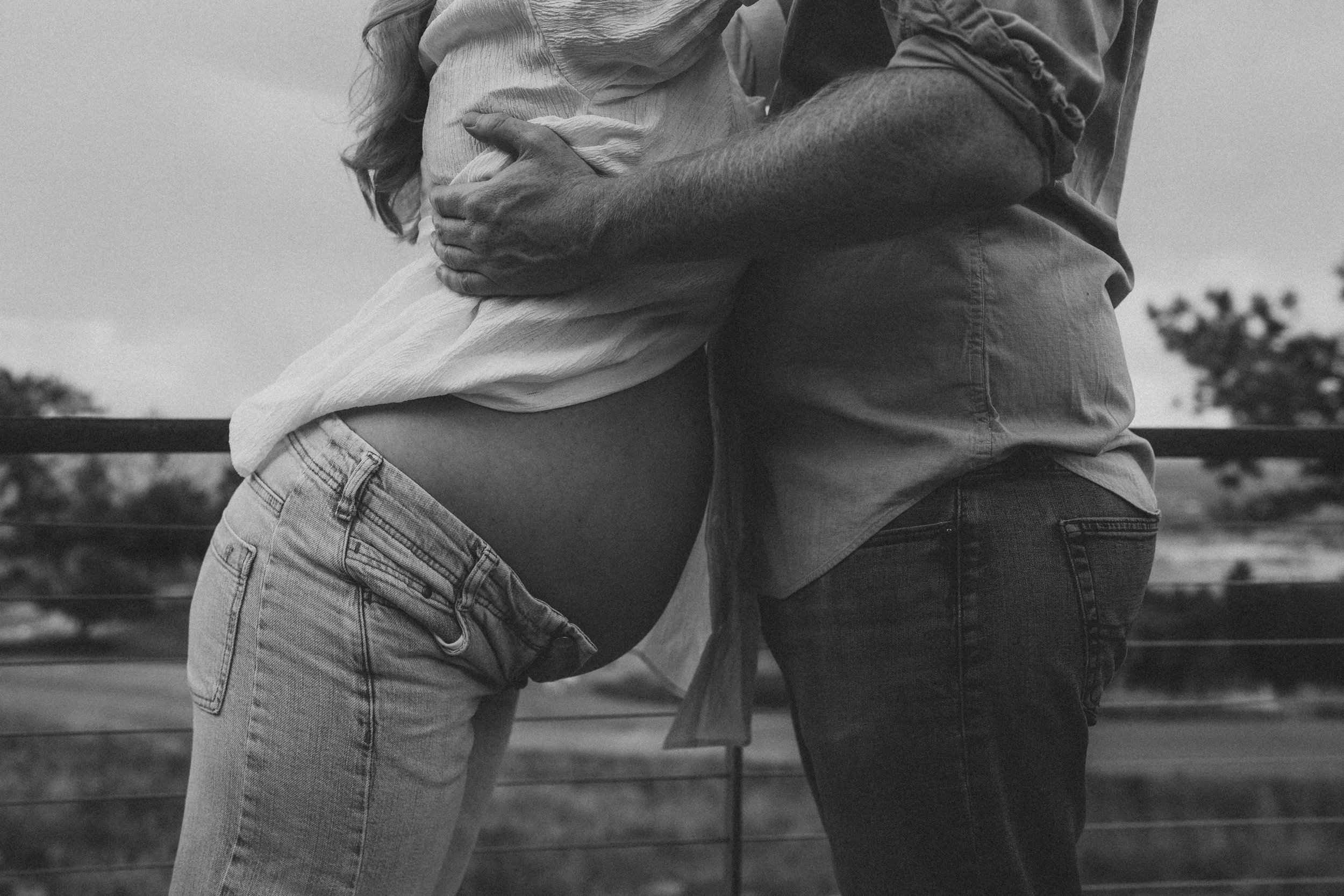 A black-and-white photo of a couple hugging outdoors, showing only their torsos and arms. The woman is wearing jeans and a light-colored top, while the man is in dark jeans and a button-up shirt. Their embrace is close, with their arms wrapped around