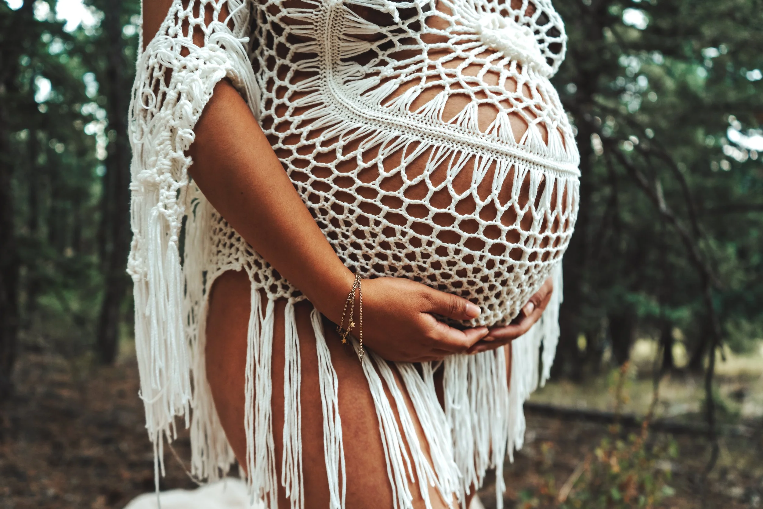 A pregnant person holding their belly outdoors, wearing a white, woven, open-knit dress or cover-up in a forest setting.