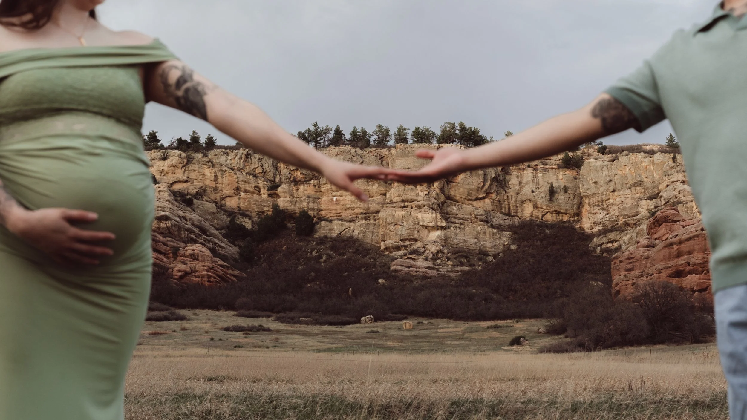 A pregnant woman in a green dress and a man are holding hands outdoors with rocky cliffs and trees in the background.