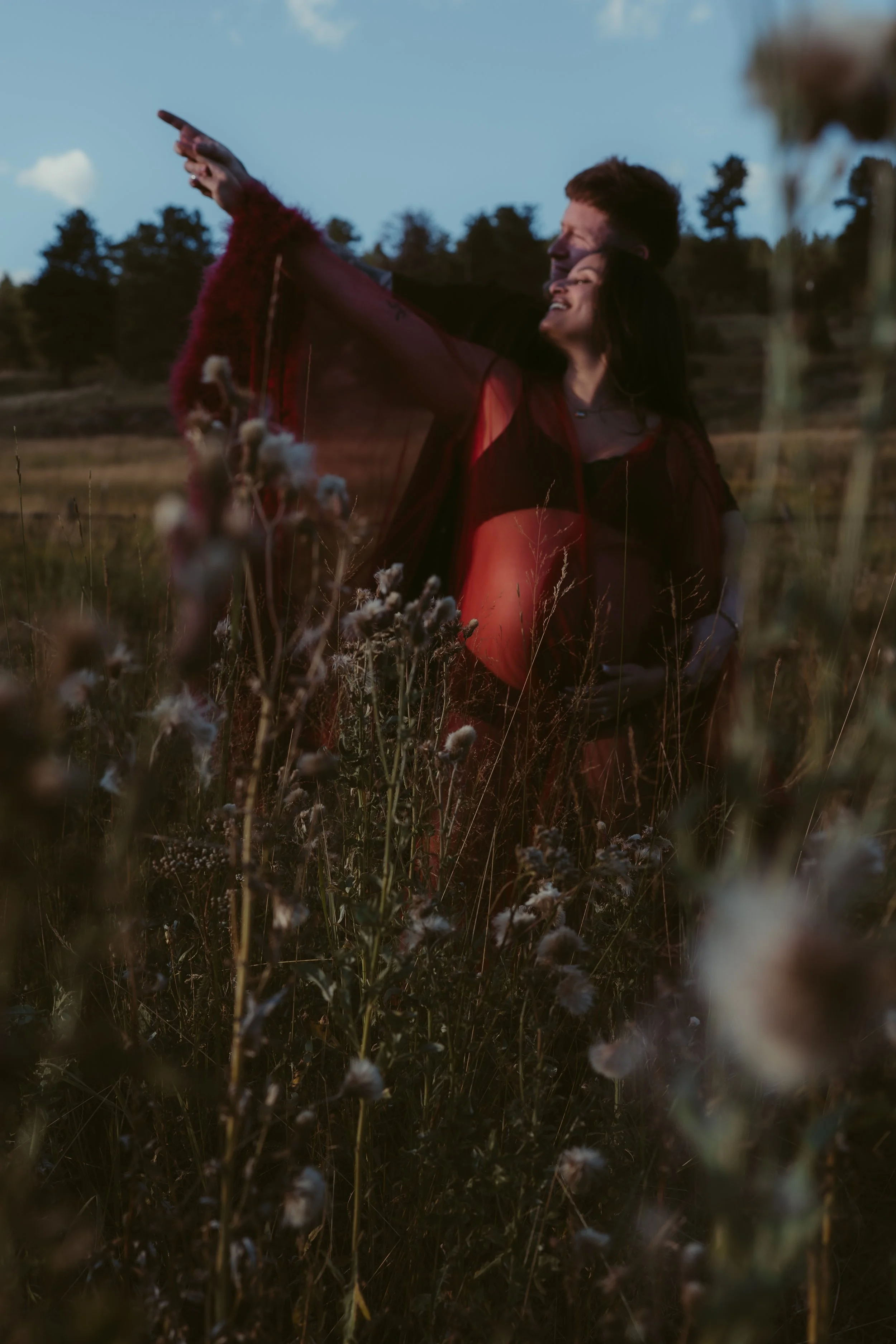 A couple in a field of tall grass and wildflowers, smiling and embracing as they point into the distance during sunset or dusk.
