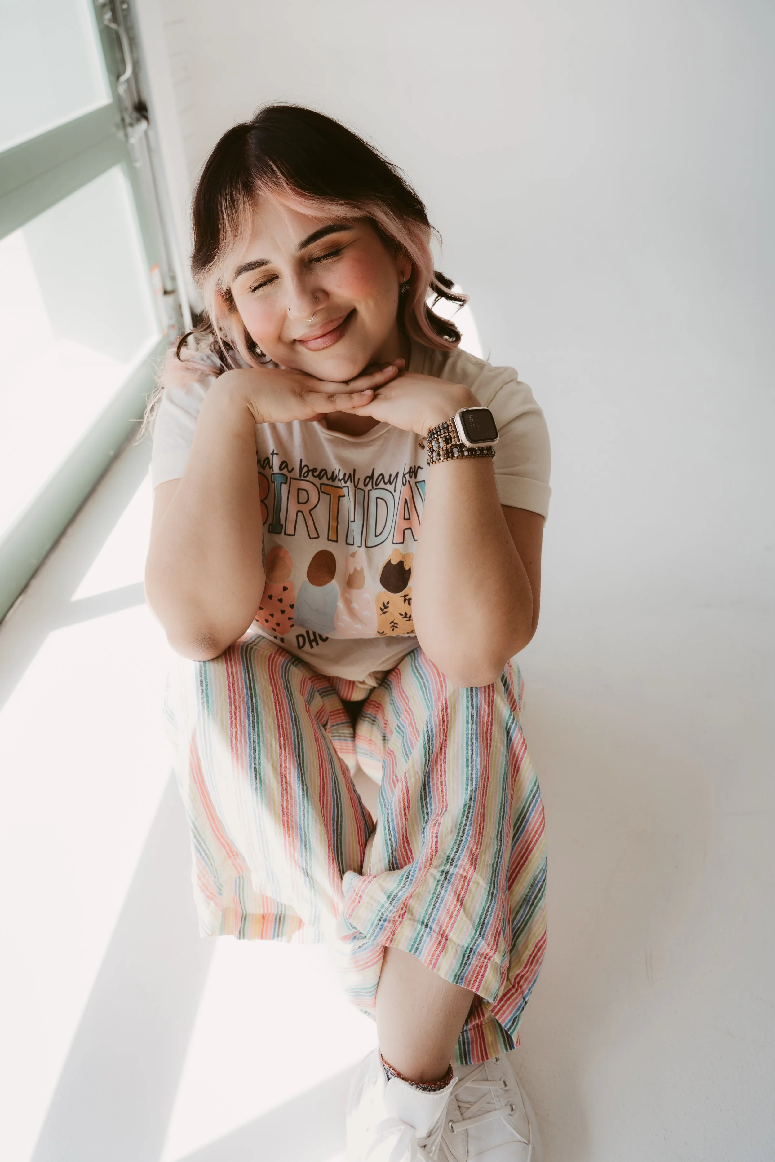 A young woman with pink and black hair, smiling with her eyes closed, resting her chin on her hands. She is wearing a birthday-themed T-shirt, a striped skirt, white sneakers, and a smartwatch. She is standing in a bright room with natural light coming through a window.