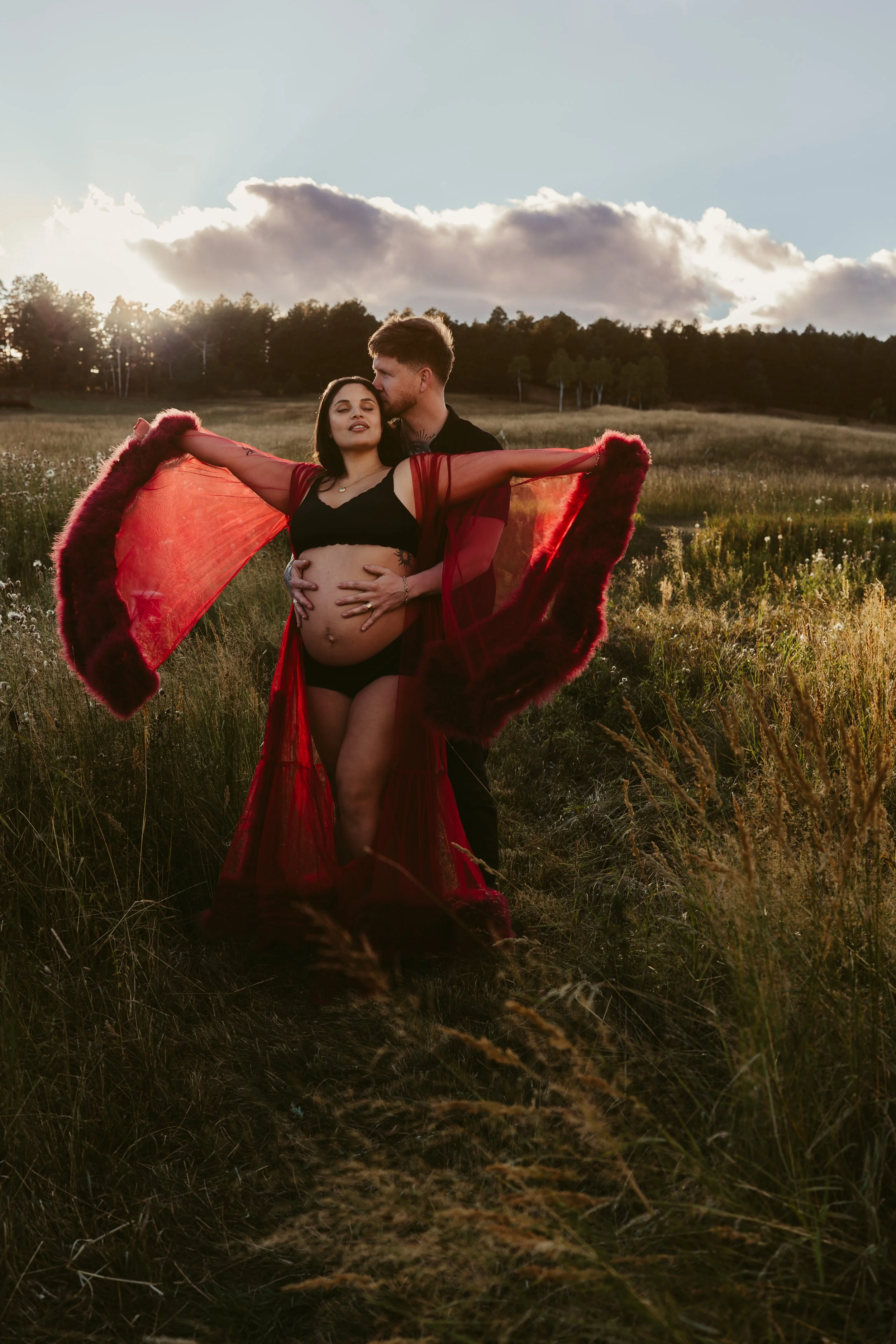 A couple standing in a grassy field at sunset. The woman, who appears pregnant, has her eyes closed and is leaning into the man. She is wearing a black crop top and a flowing red and black shawl with fur trim. The man is holding her with one hand on 