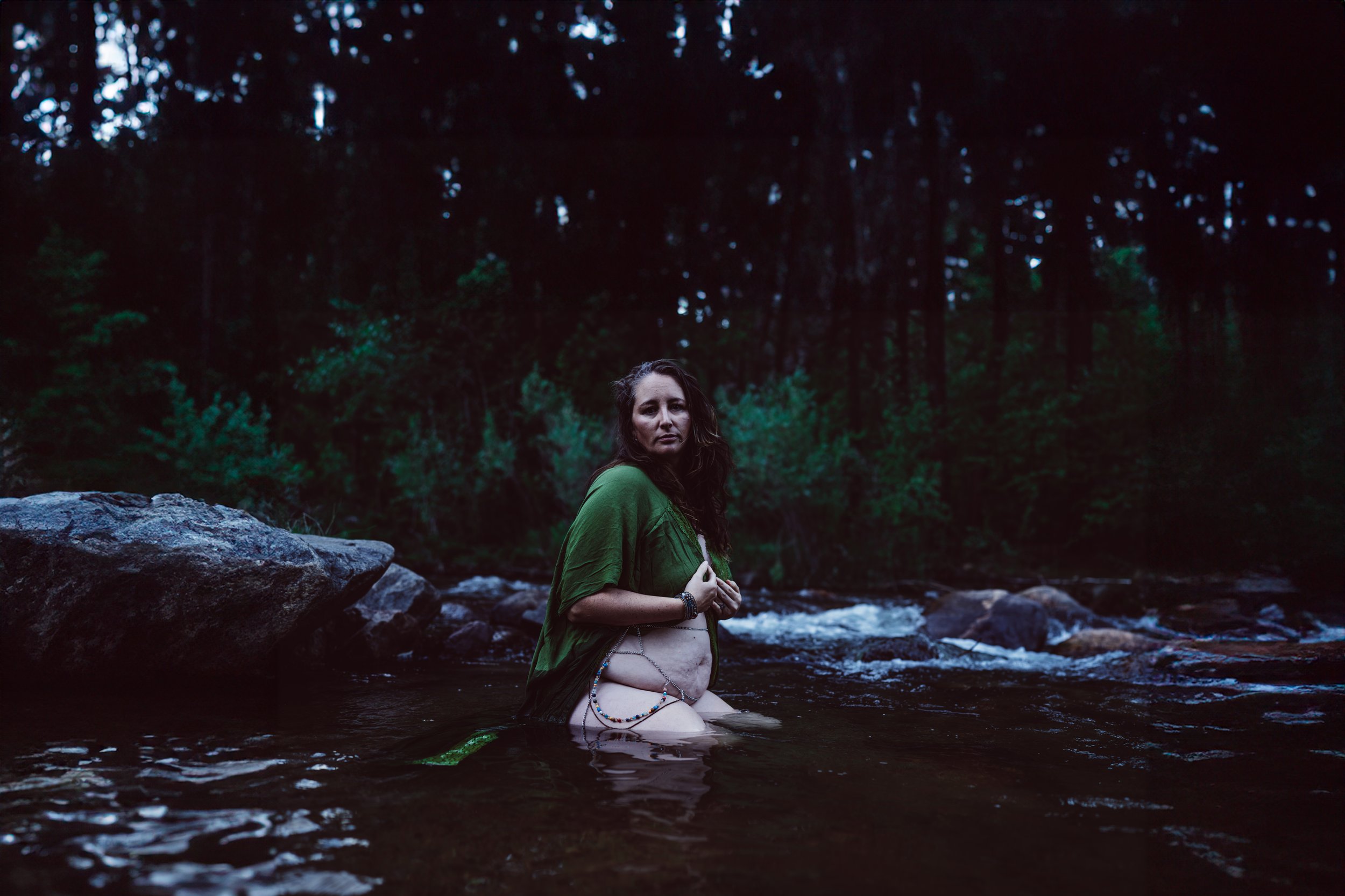 A woman with long dark hair standing in a river, wearing a green top and decorated with jewelry, surrounded by a forested area.