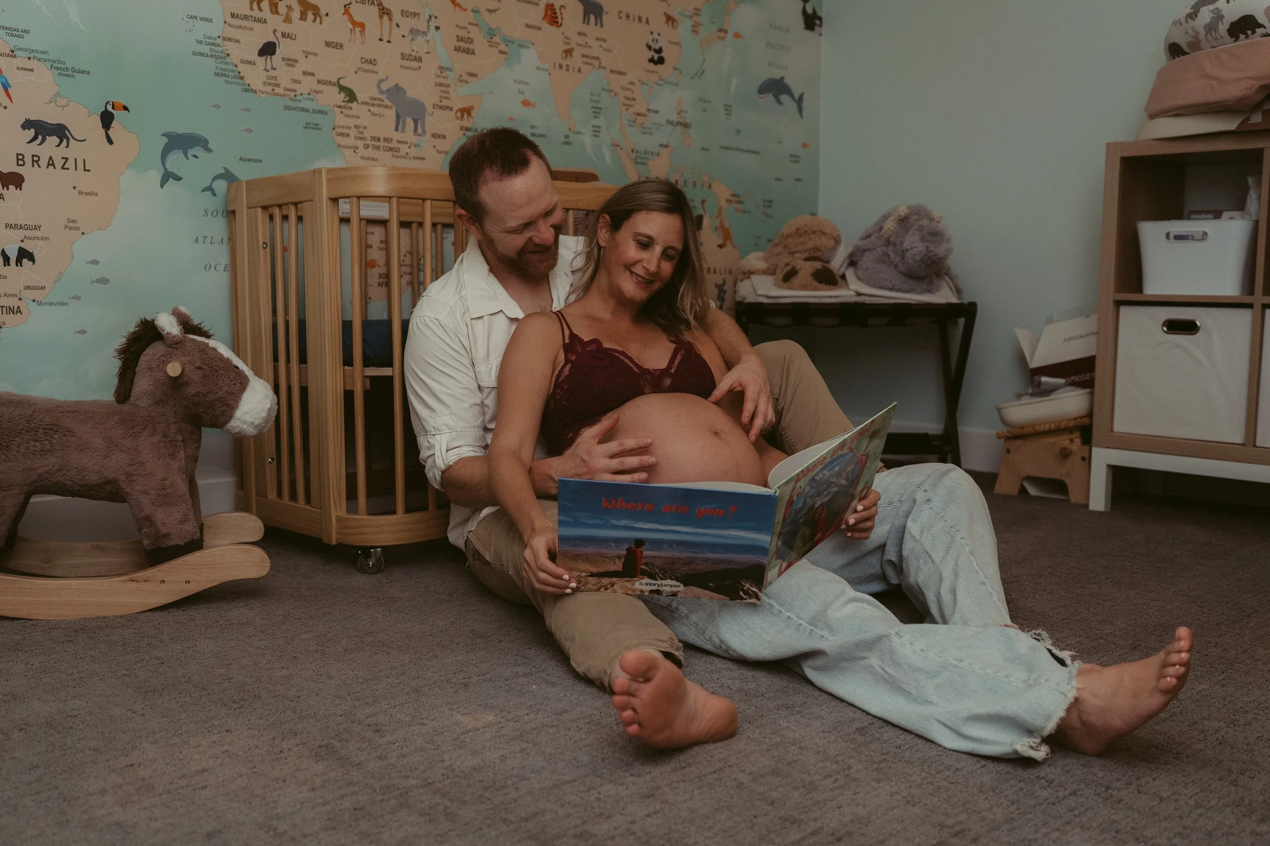 A man and woman sitting on the floor in a nursery room, with a world map wall in the background. The woman is pregnant, holding a children's book titled 'Where are you?' while the man embraces her. There is a plush horse toy on a wooden rocking horse