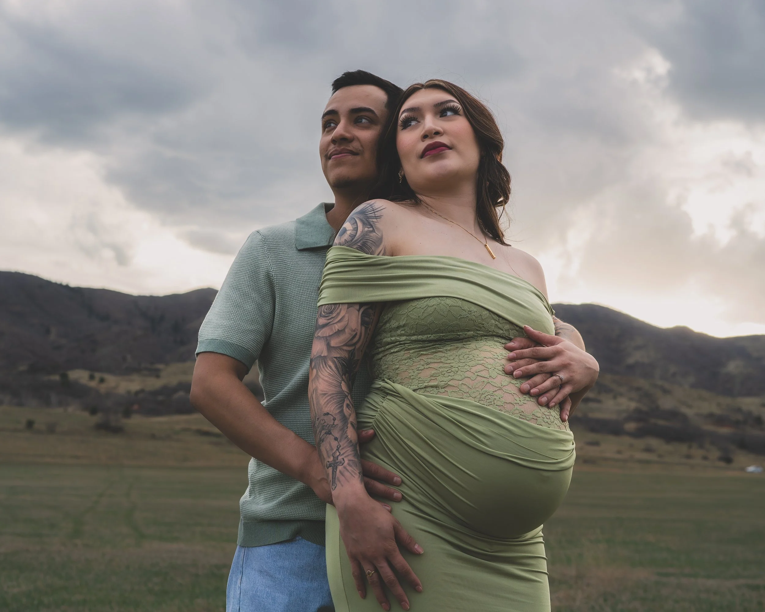 A pregnant woman wearing a green dress and a man standing close behind her, both outdoors with cloudy sky and mountains in the background.
