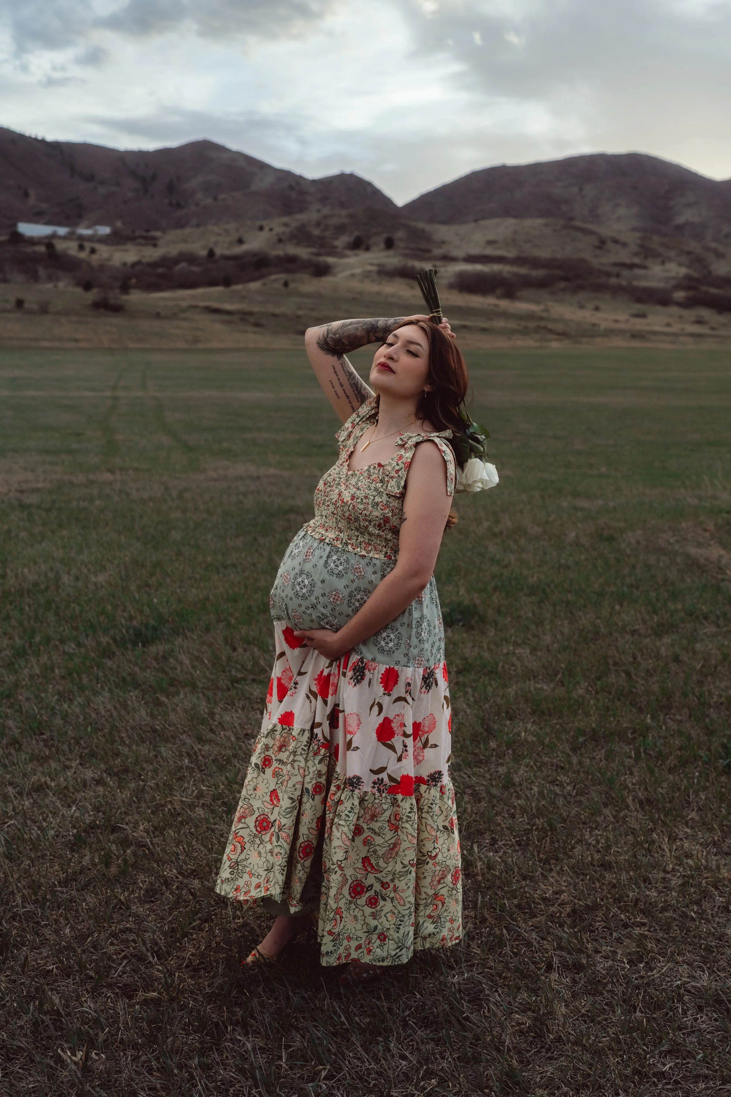 A pregnant woman with red hair and tattoos on her arm, wearing a floral dress, standing in a field with hills and cloudy sky in the background, holding white roses behind her head with one hand and her belly with the other.