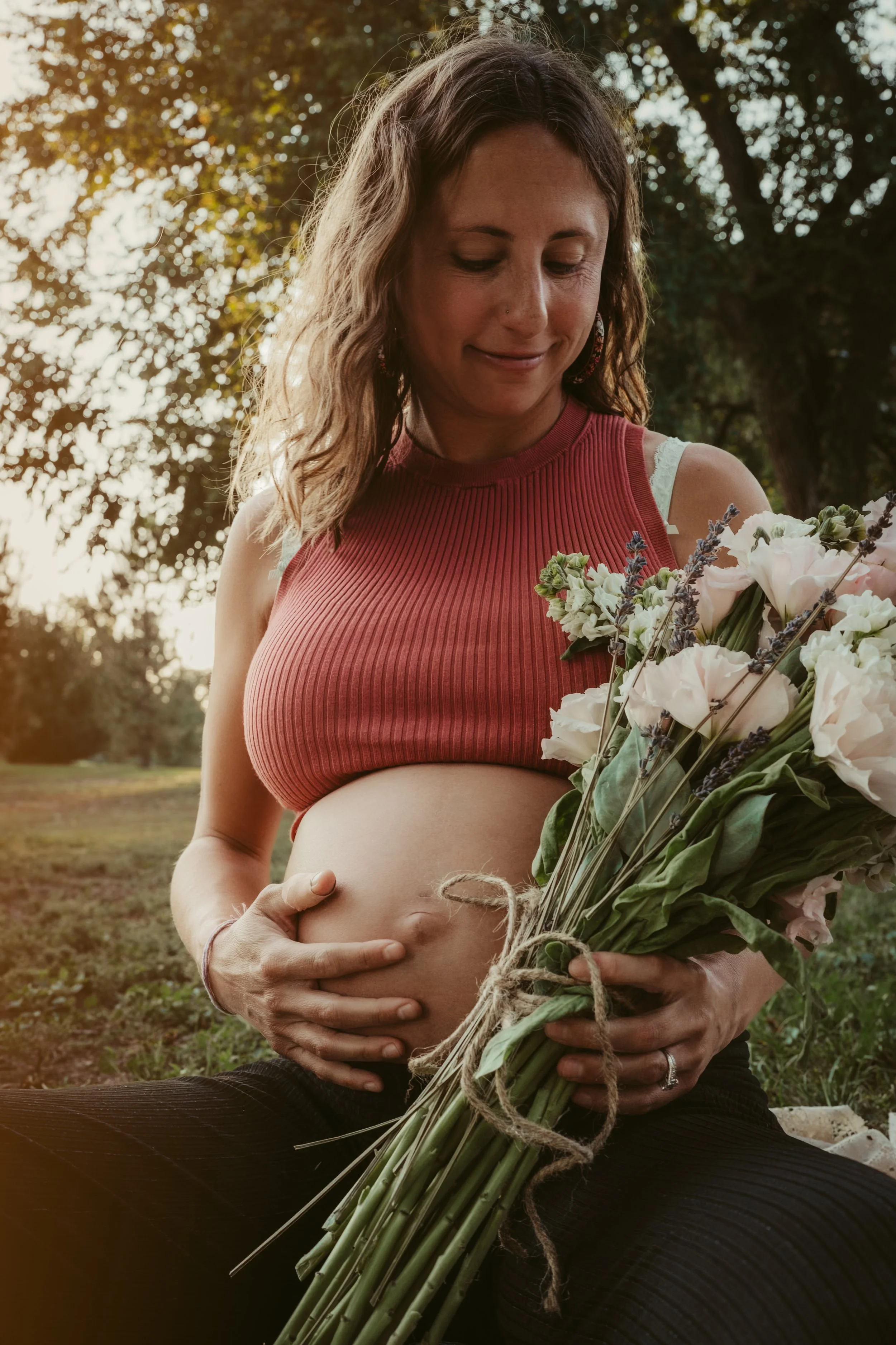 A pregnant woman sitting outdoors holding a bouquet of flowers, with her hands gently resting on her belly, during golden hour.