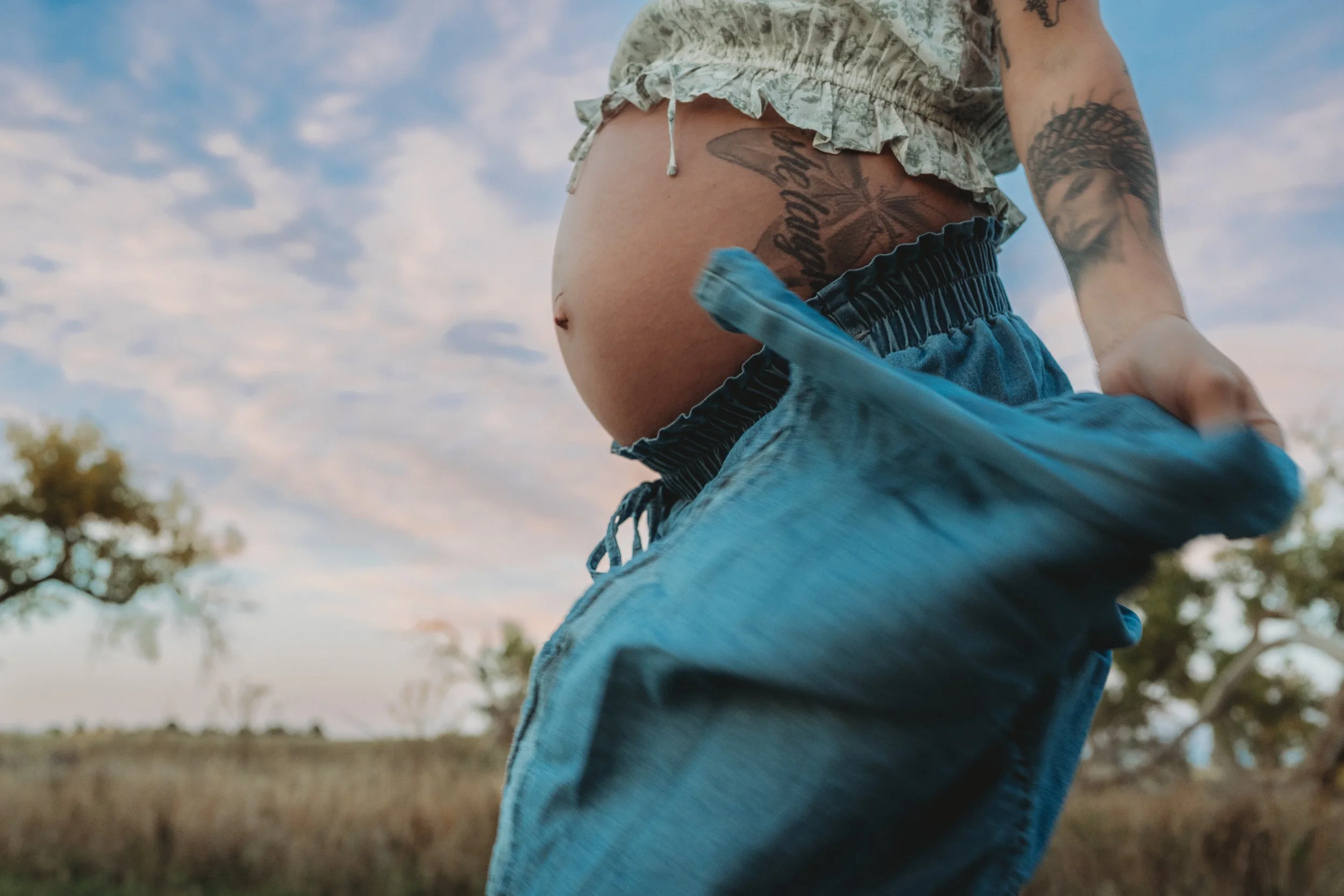 A person in blue jeans and a light-colored shirt with floral ruffles, displaying tattoos on their torso and arm, standing outdoors under a partly cloudy sky, with trees and open fields in the background.