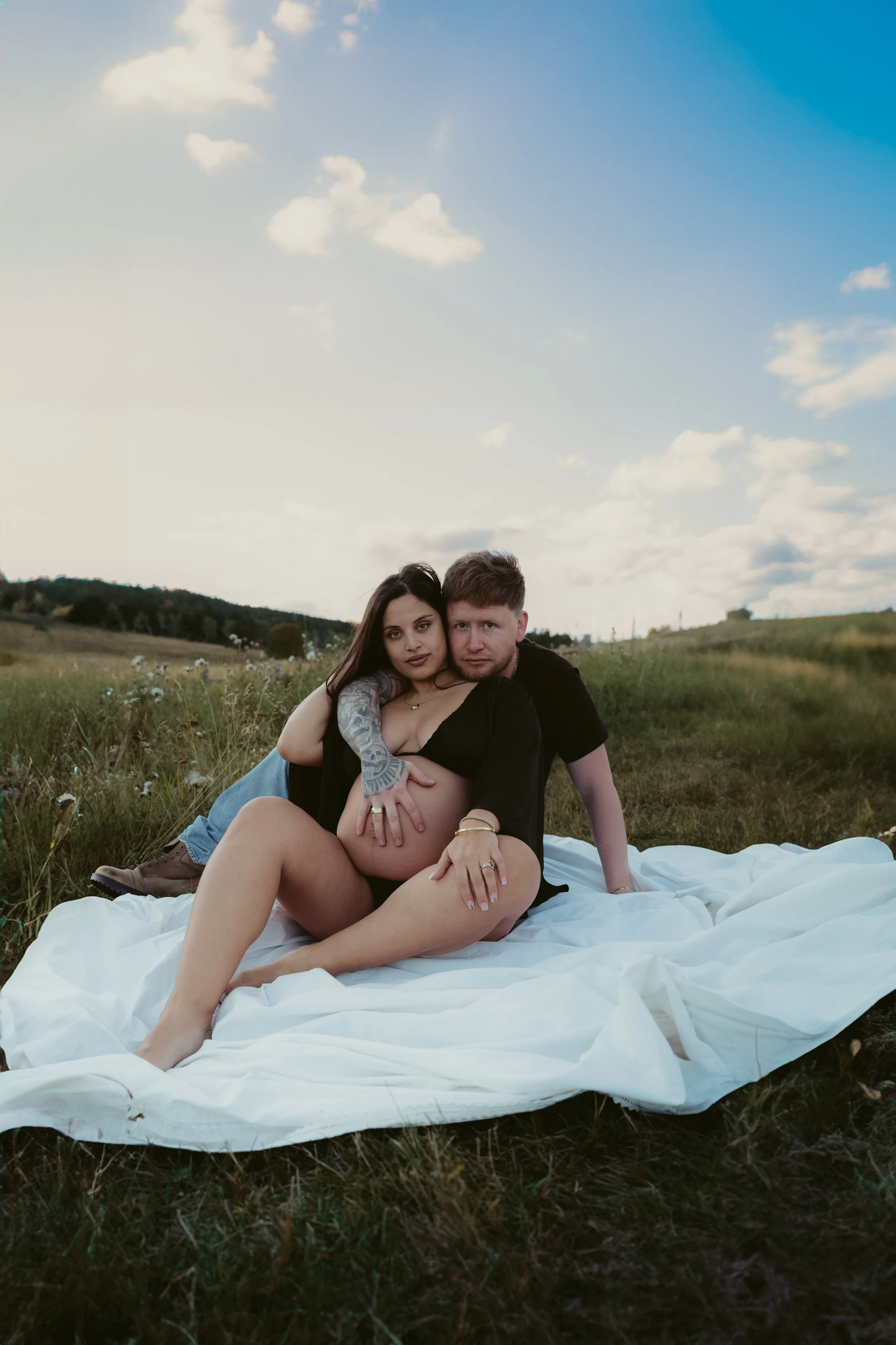 A couple sitting on a white blanket in a field, with a woman appearing pregnant, the man behind her with his arms around her. The woman has dark hair and tattoos on her arm, and the man has short hair. The scene is outdoors with a cloudy sky and open