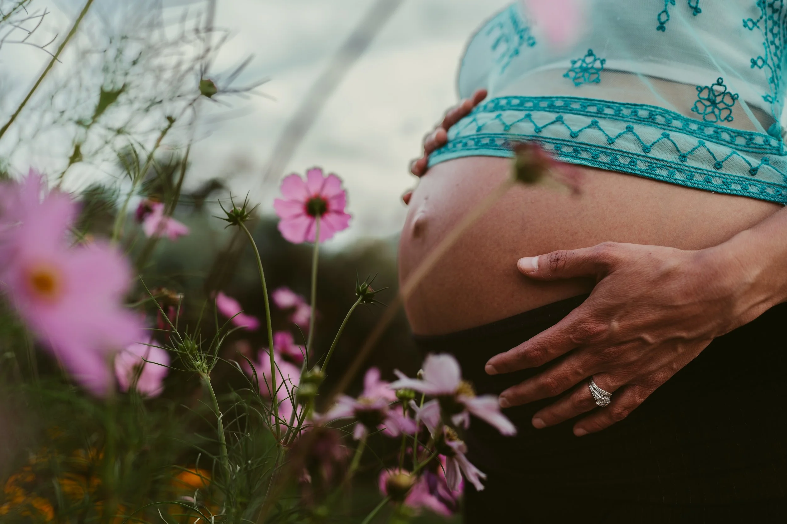 Close-up of a pregnant woman holding her belly outdoors with pink flowers in the foreground and blue embroidered top.