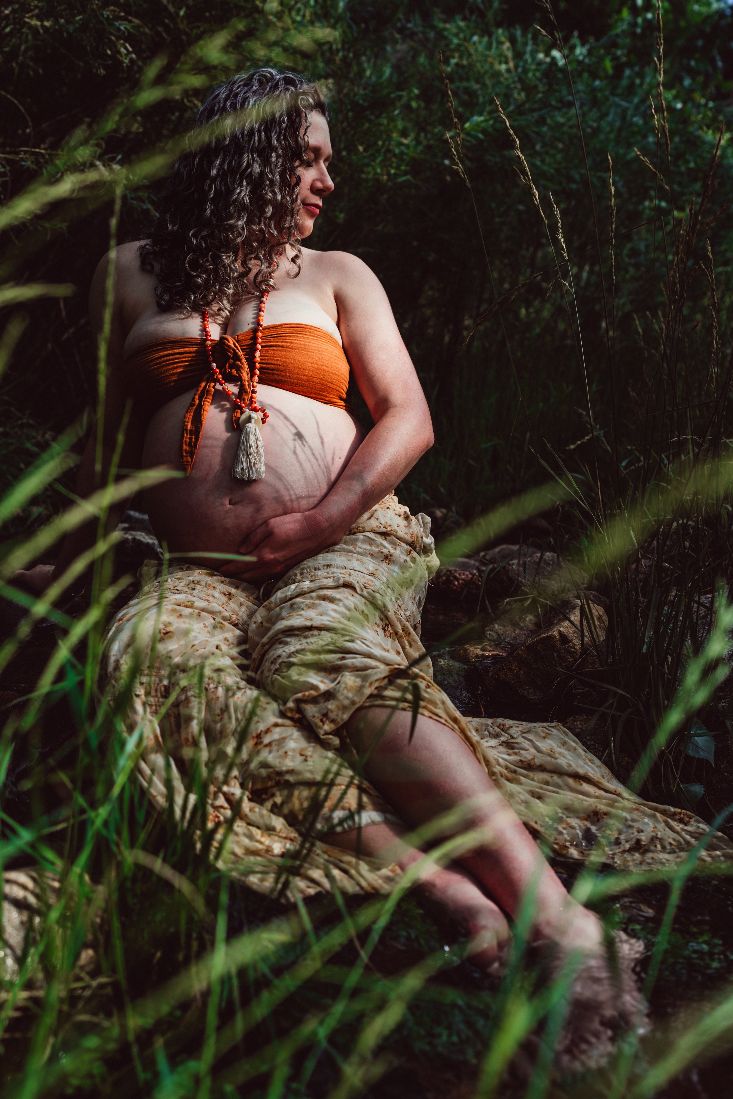 A woman with curly hair and light skin sitting in a natural outdoor setting surrounded by tall grass and greenery, wearing a strapless orange top and patterned beige pants. She is looking to the side, with her hand on her stomach.