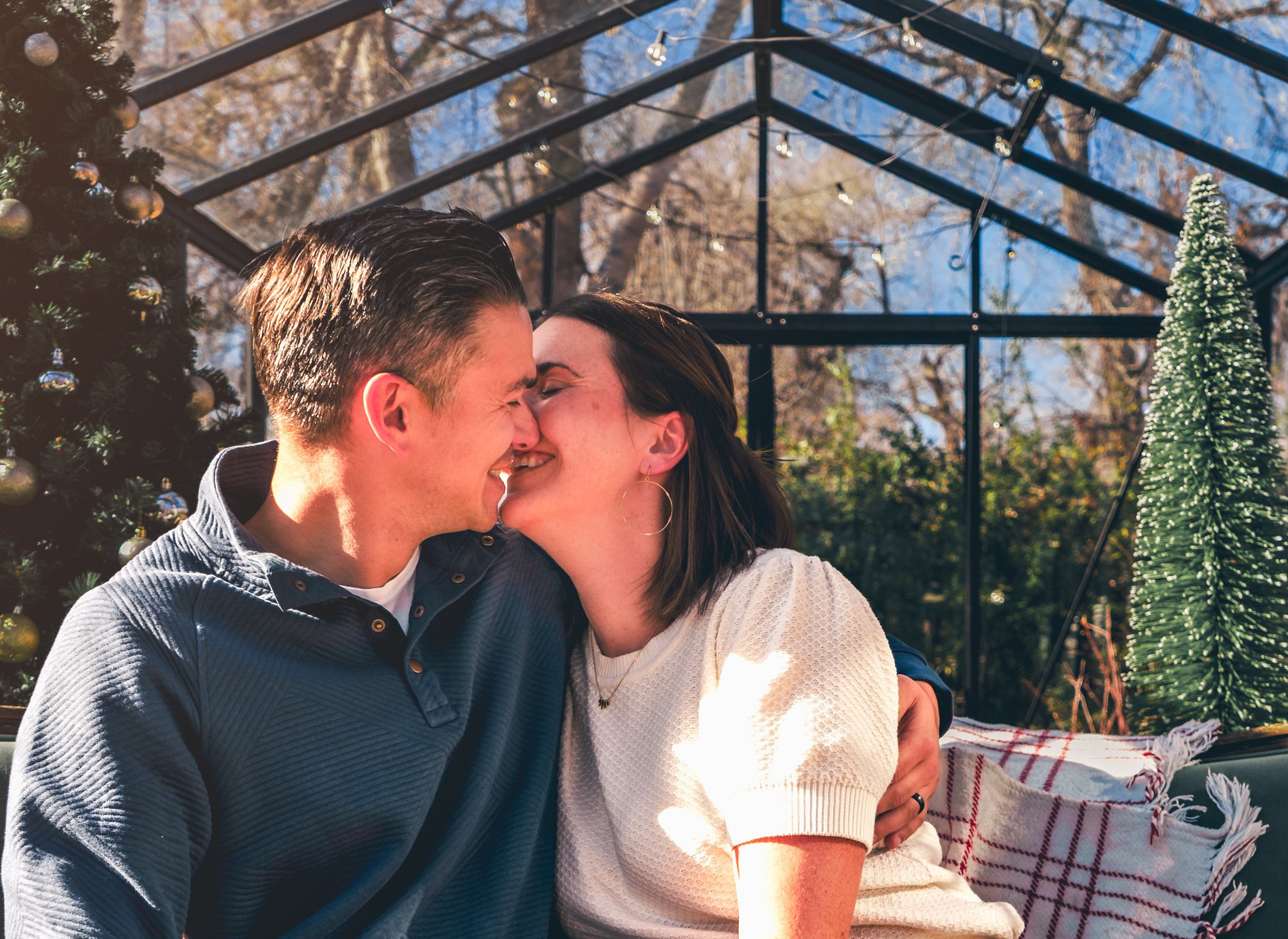 A couple smiling and embracing in a holiday-themed greenhouse, with a decorated Christmas tree and string lights in the background.