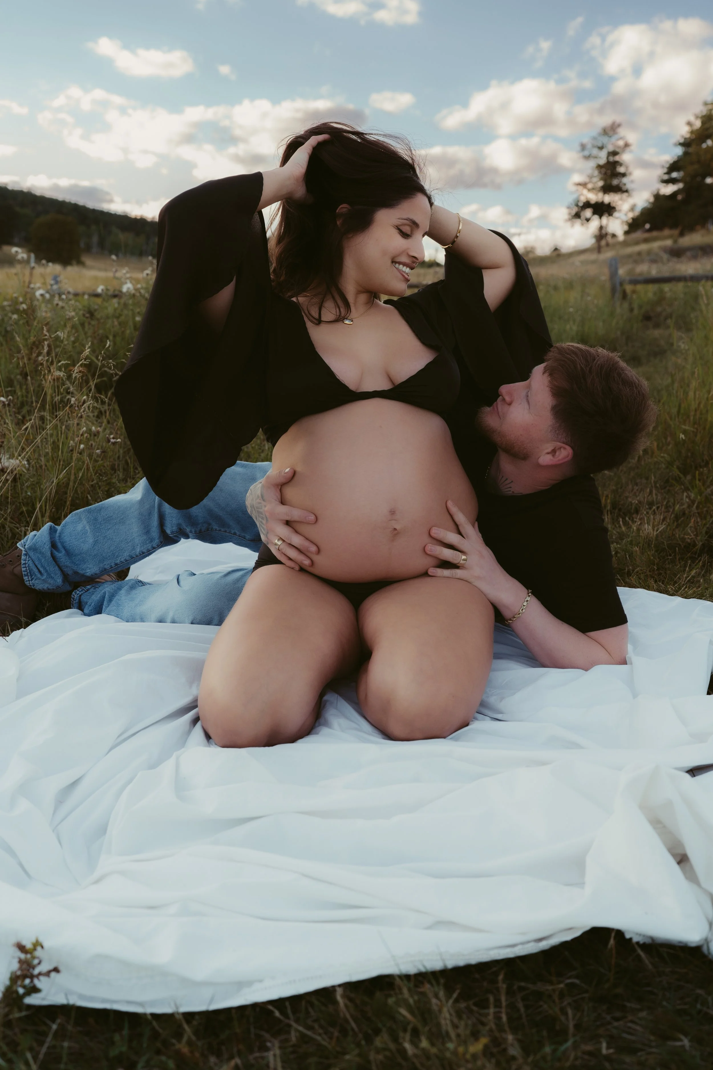 A pregnant woman and a man are sitting on a white blanket in the grass outdoors, sharing a joyful moment as they look at each other, with the woman lifting her hair and the man looking lovingly at her.