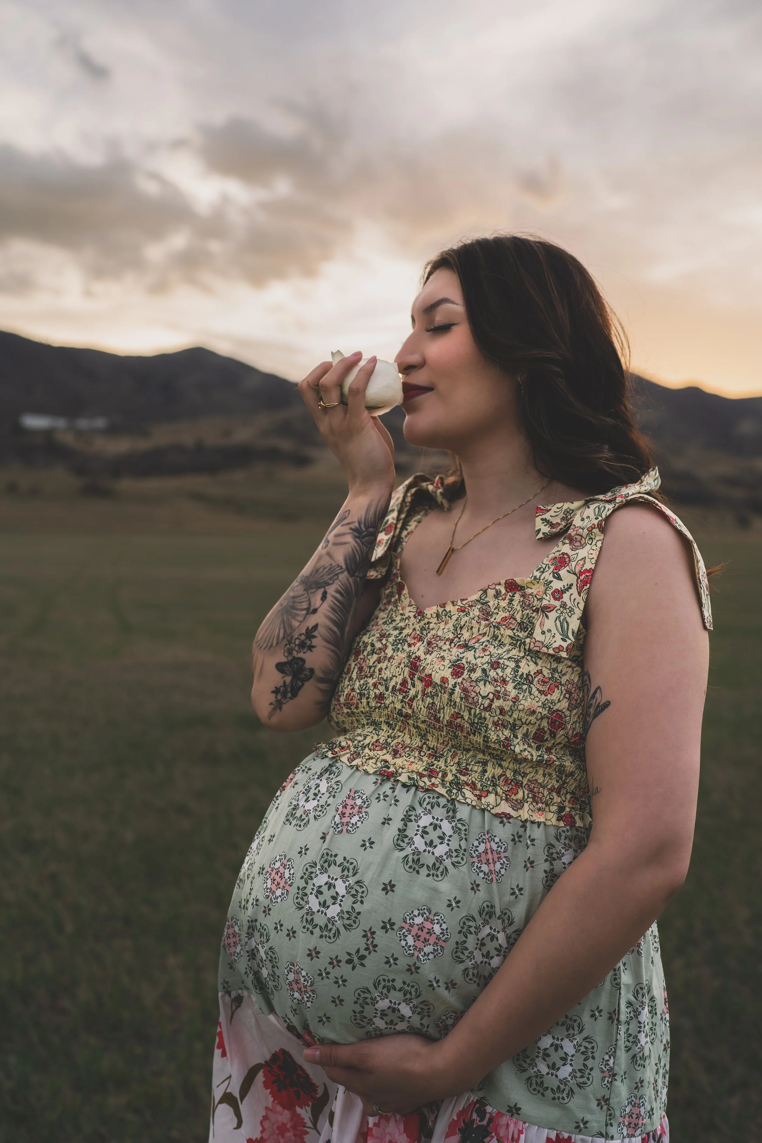 A woman with dark wavy hair and tattoos on her arm, wearing a floral dress, is holding a flower in front of her nose with her eyes closed, standing outdoors during sunset in a grassy field with mountains in the background.