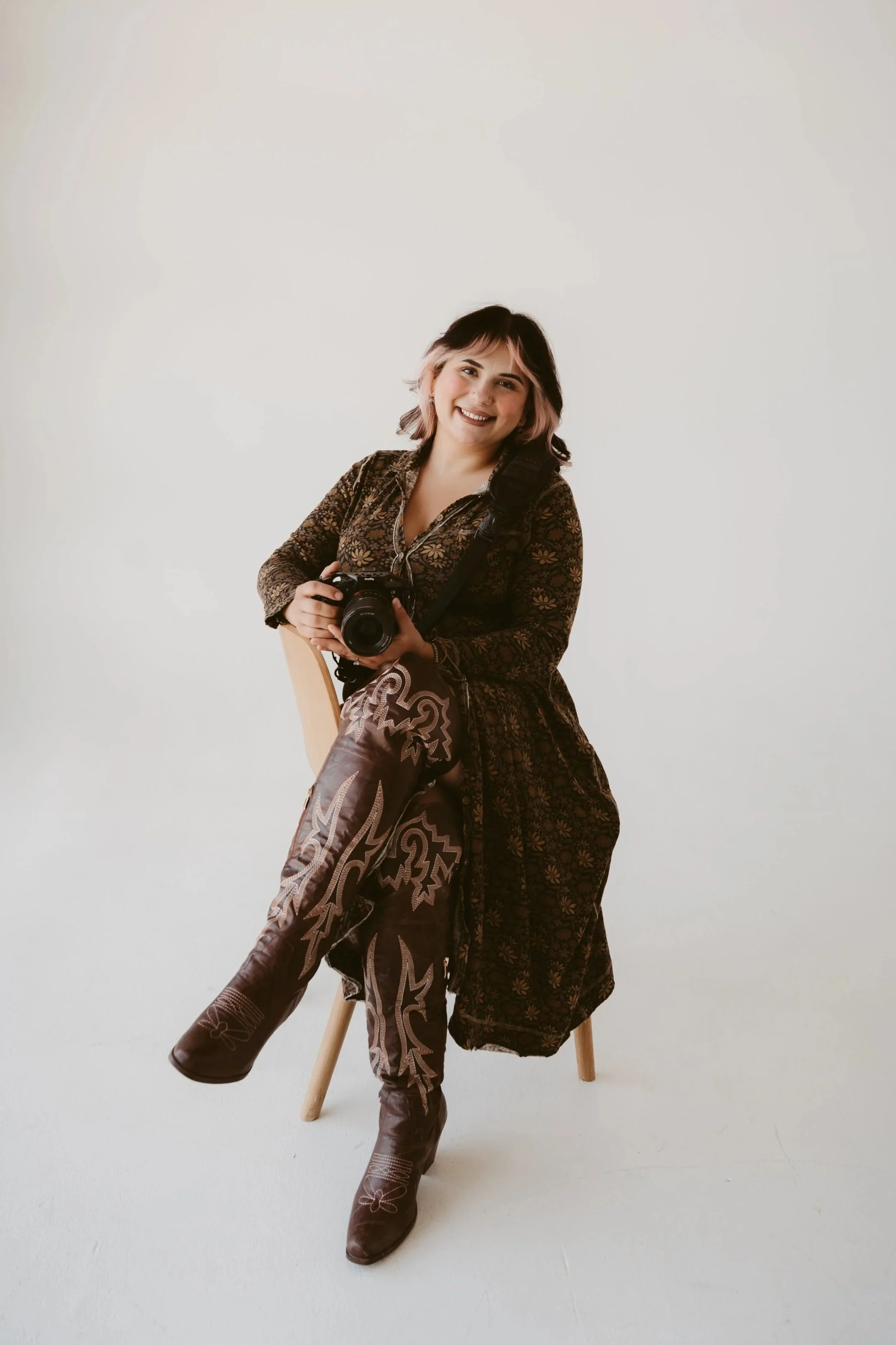 A woman sitting on a wooden stool against a plain white background, smiling, holding a camera, wearing patterned brown western-style shirt, matching patterned brown pants, and brown cowboy boots with white embroidery.
