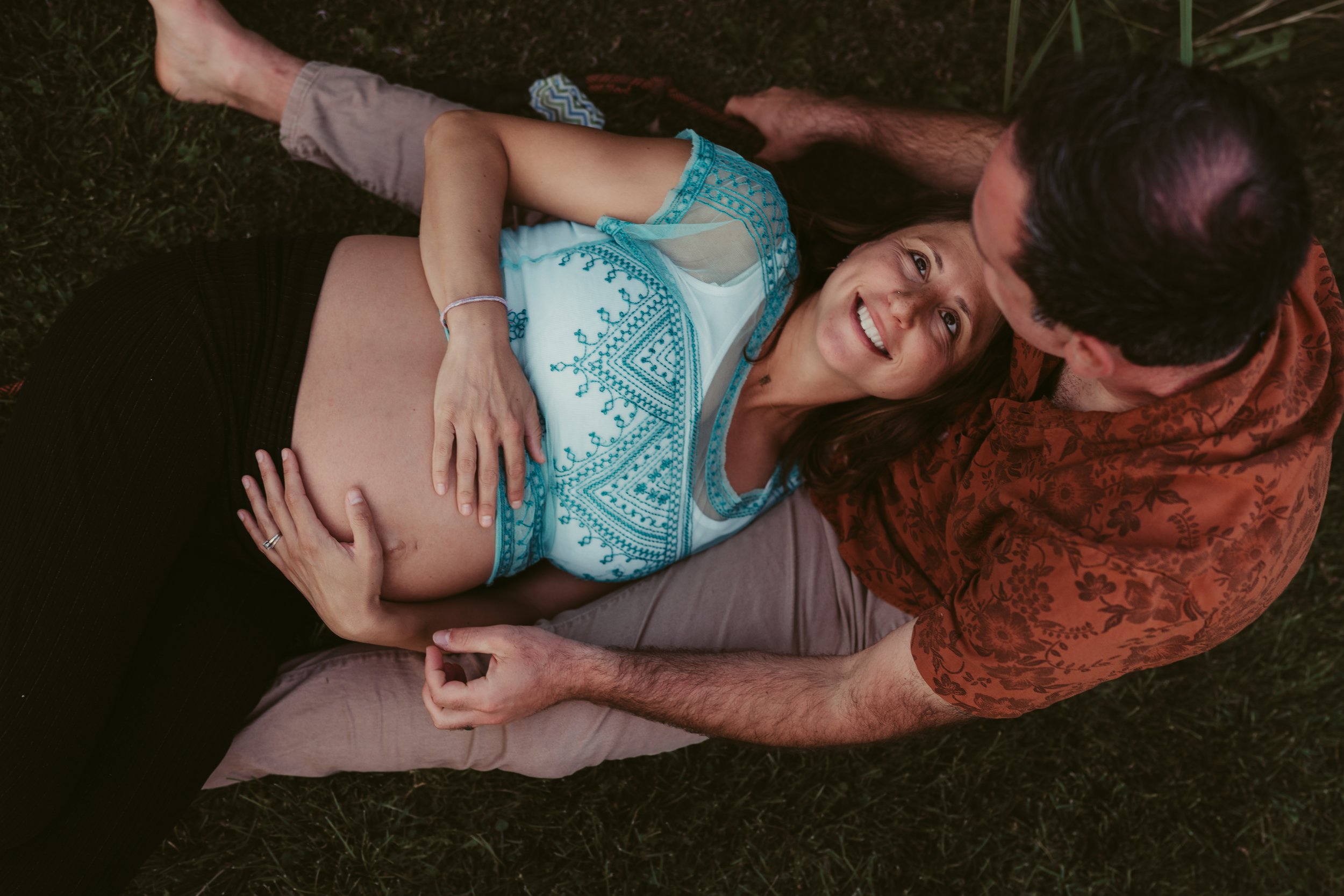 A pregnant woman lying on A man's lap outdoors, smiling and looking at him, with green grass around them.
