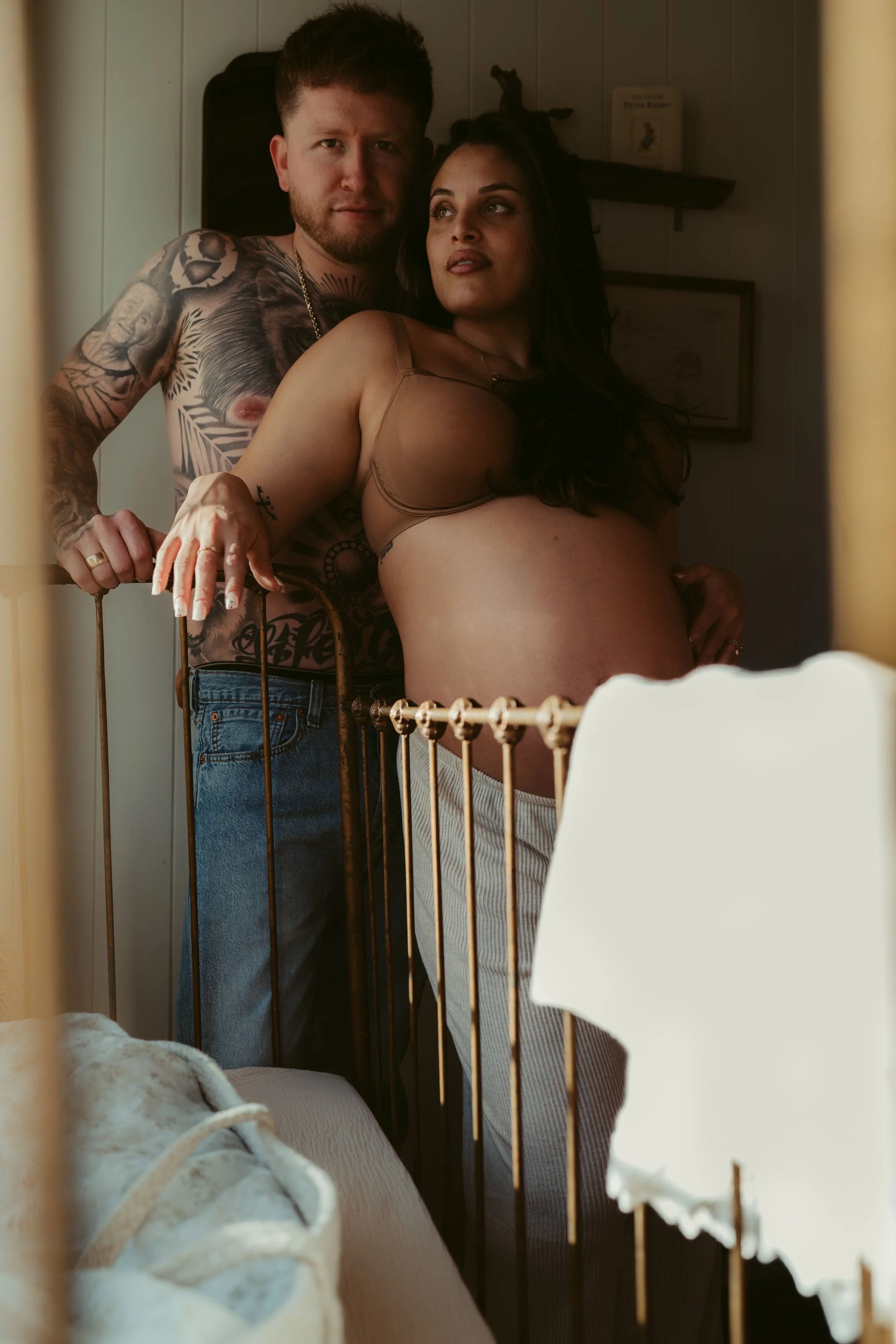 A couple in a bedroom, the man with tattoos and the woman pregnant, standing behind a bed with white bedding, seen through a yellow bed rail.