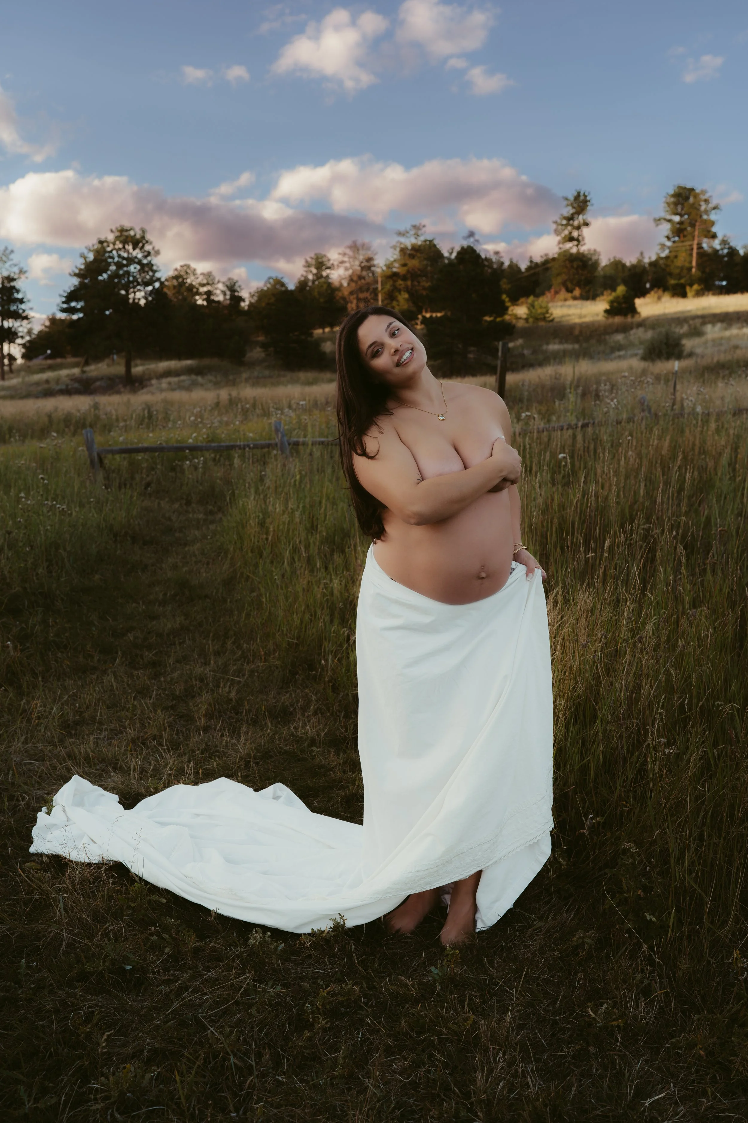 A pregnant woman stands in a grassy field with a white cloth draped around her lower body. She is topless, covering herself modestly with one arm, and smiling at the camera. The background includes trees, a fence, and a sky with clouds.