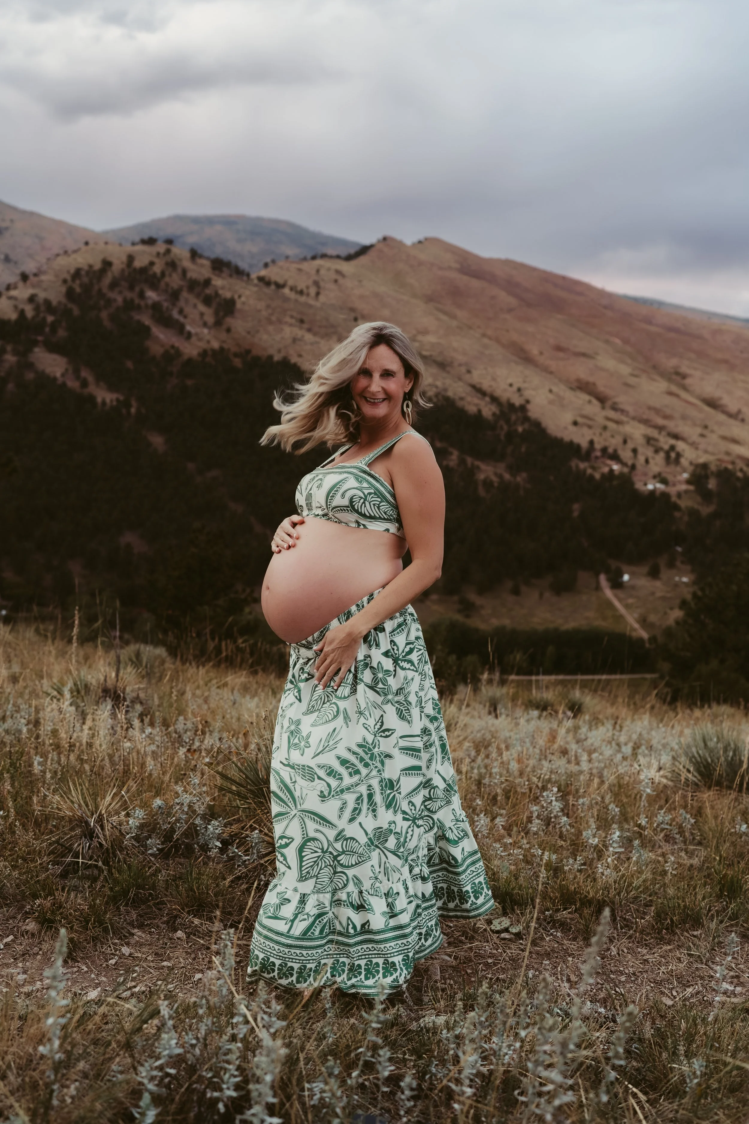 A pregnant woman in a green and white patterned dress standing in a field with mountains in the background