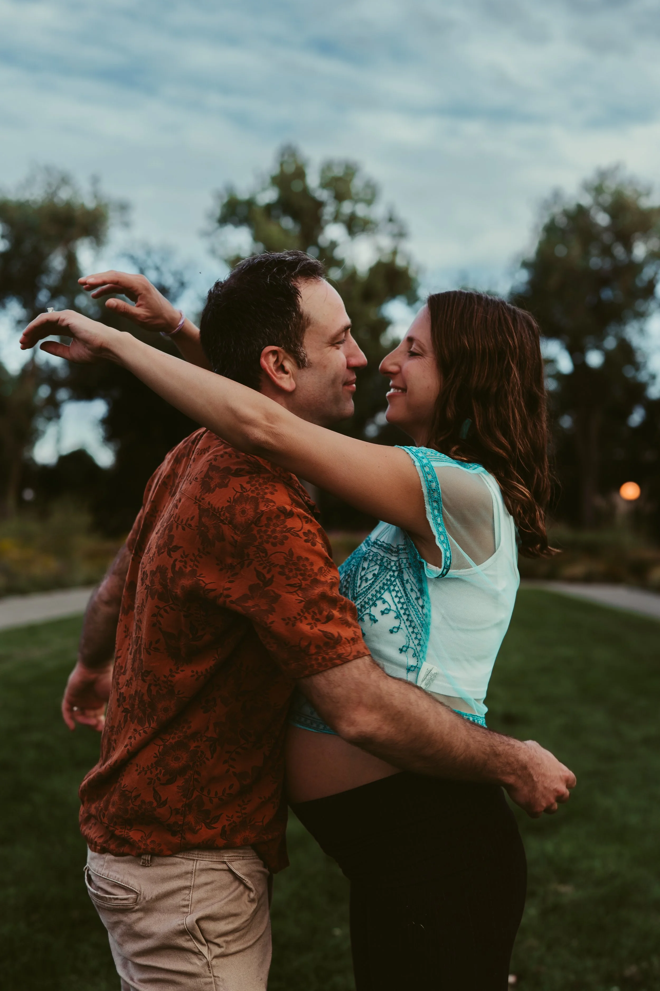 A couple dancing outdoors with arms wrapped around each other, smiling, in a park or open green space with trees and a cloudy sky.