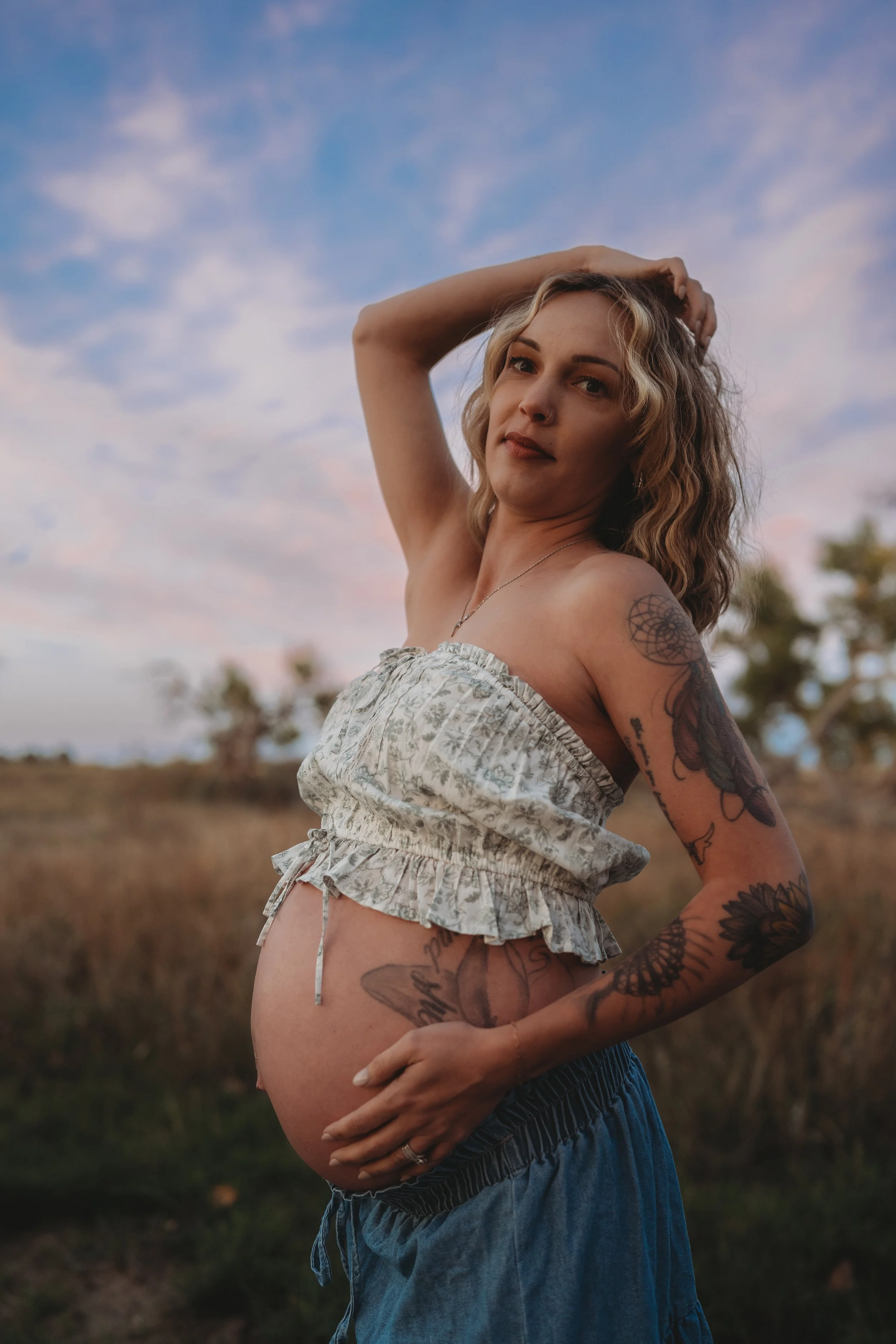A pregnant woman with tattoos on her arms and chest, wearing a floral off-shoulder top and blue skirt, standing outdoors with a sky and trees in the background, during sunset.