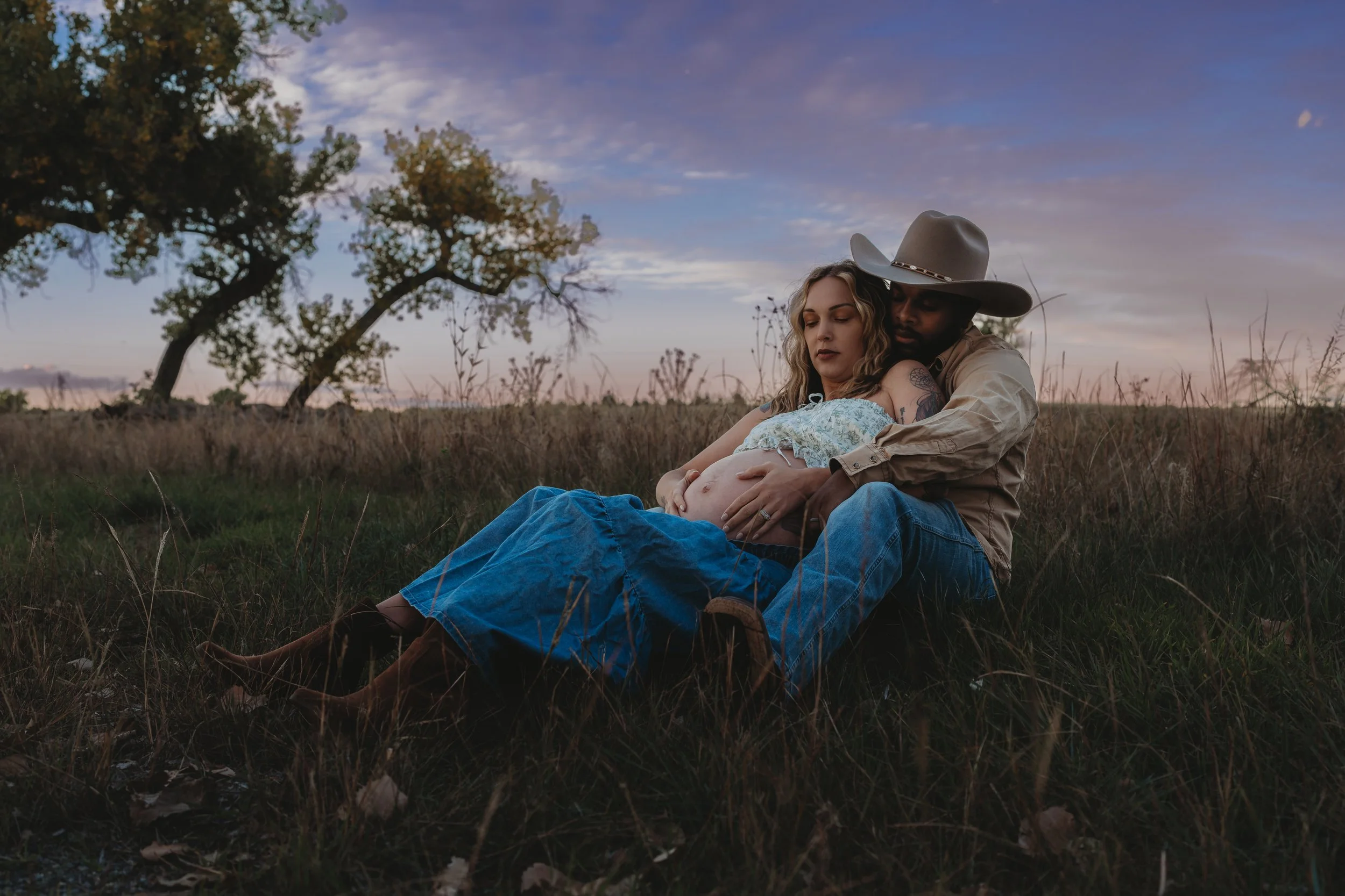 A couple sitting in a field at sunset, with the woman who appears to be pregnant and the man wearing a cowboy hat, embracing each other.