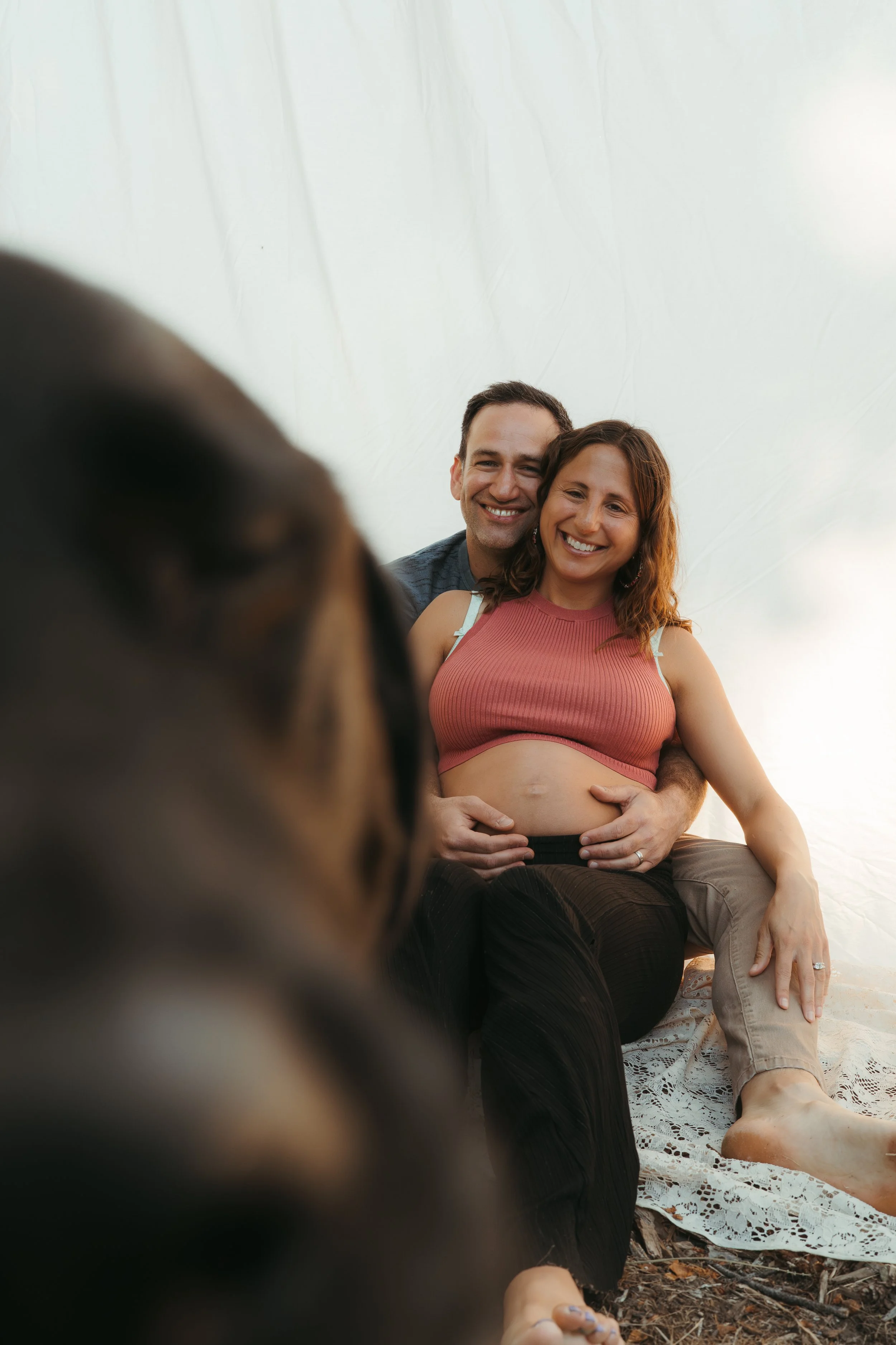 A happy couple sitting on a blanket outdoors. The woman is pregnant and smiling, with the man embracing her from behind, also smiling. The photo is taken from a low angle with a blurred foreground.